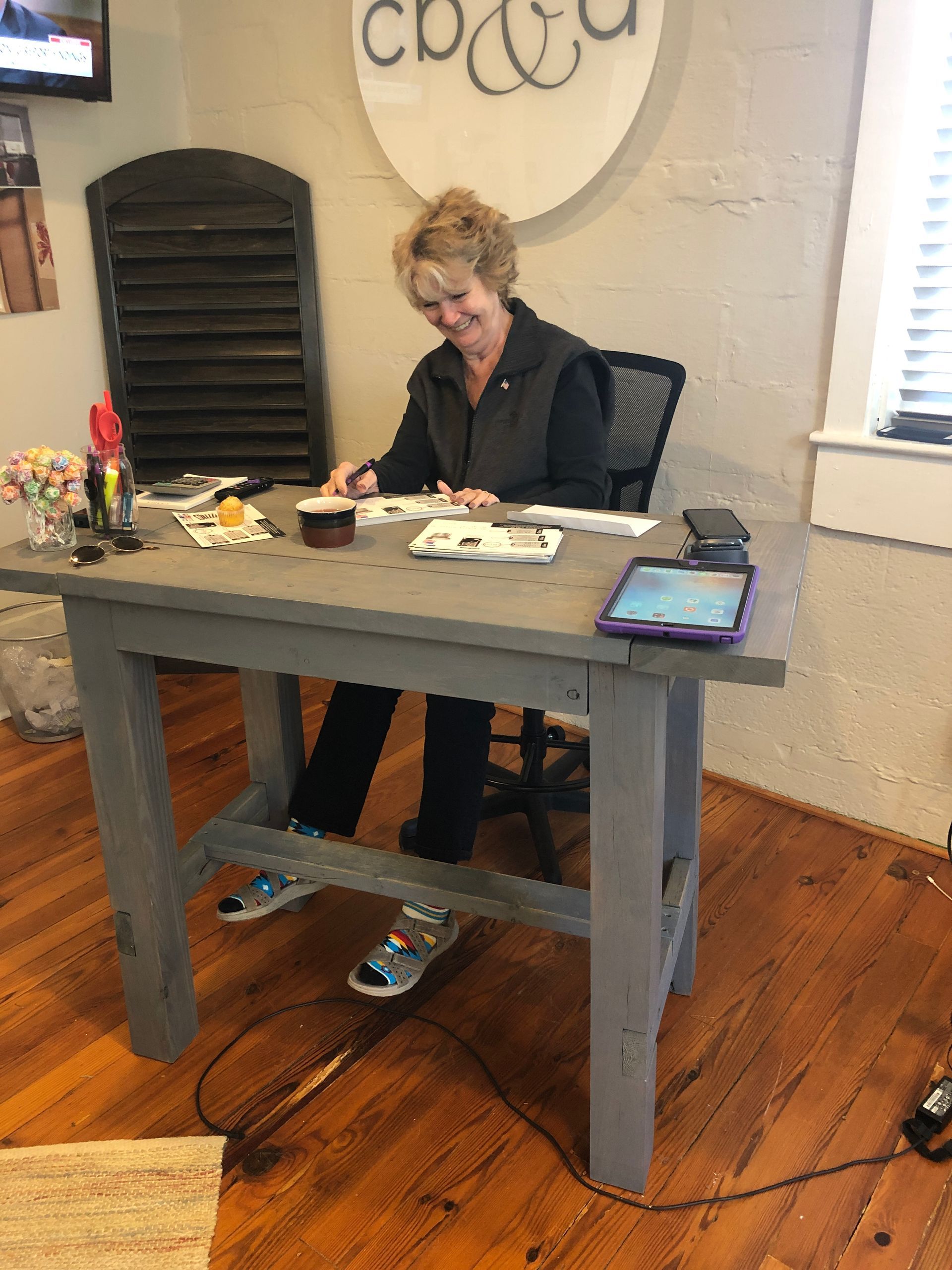 Woman sitting at a gray desk, smiling while looking at papers. Wooden floor. Sign above says 
