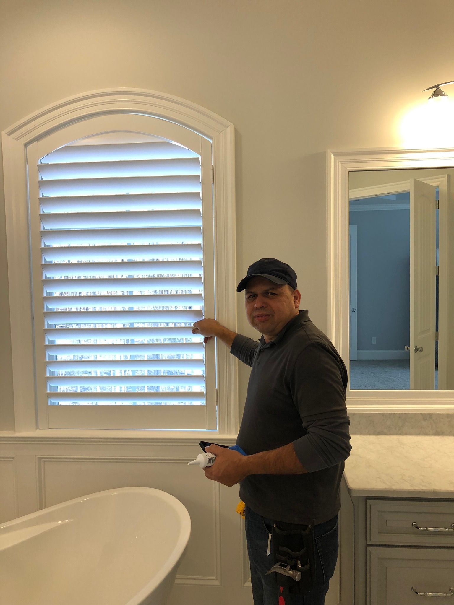 Man installing white shutters in a bathroom with a white tub and a mirror.
