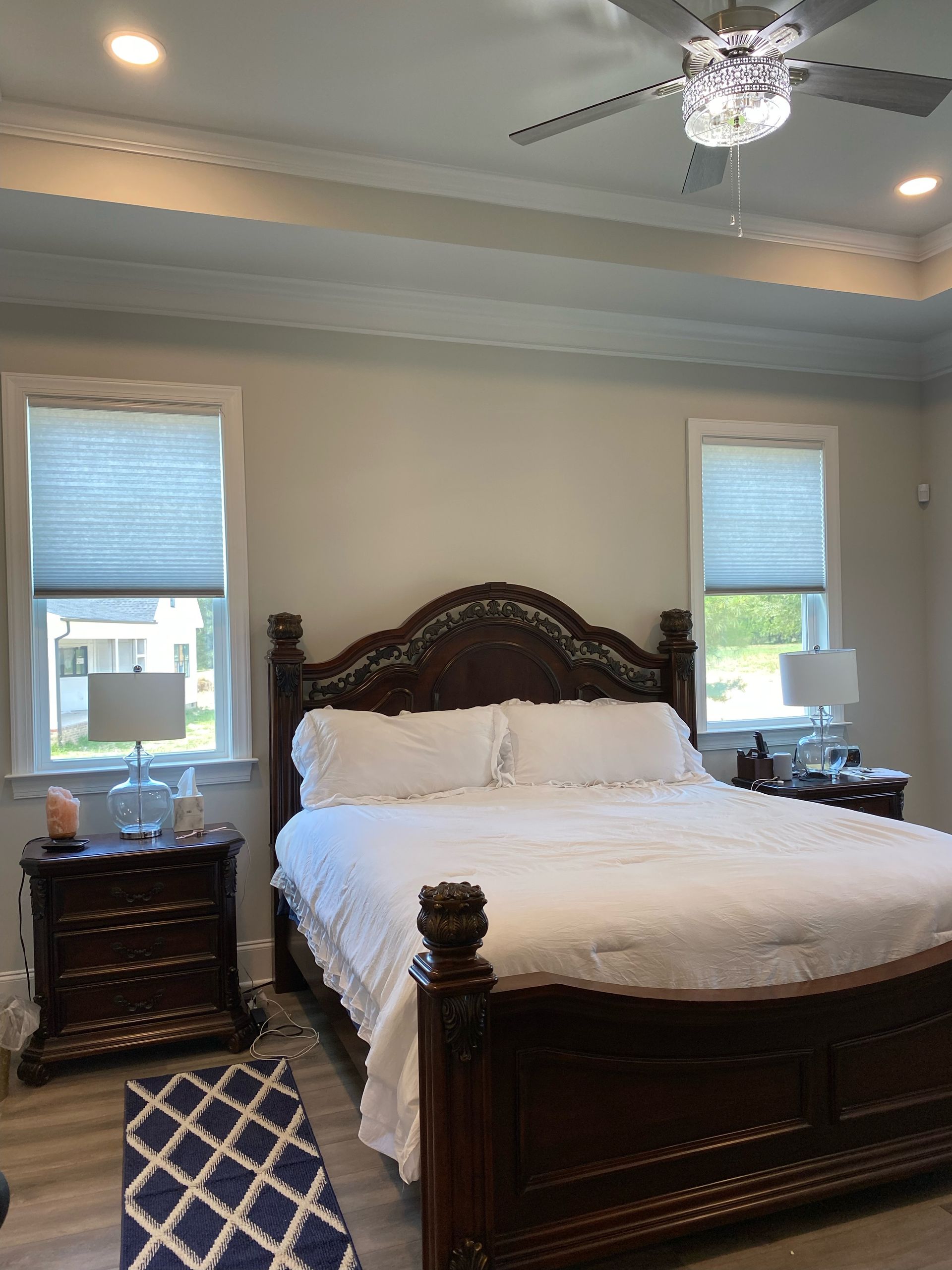 Bedroom with dark wood bed, nightstands, and blue patterned rug. Two windows with blinds and ceiling fan.