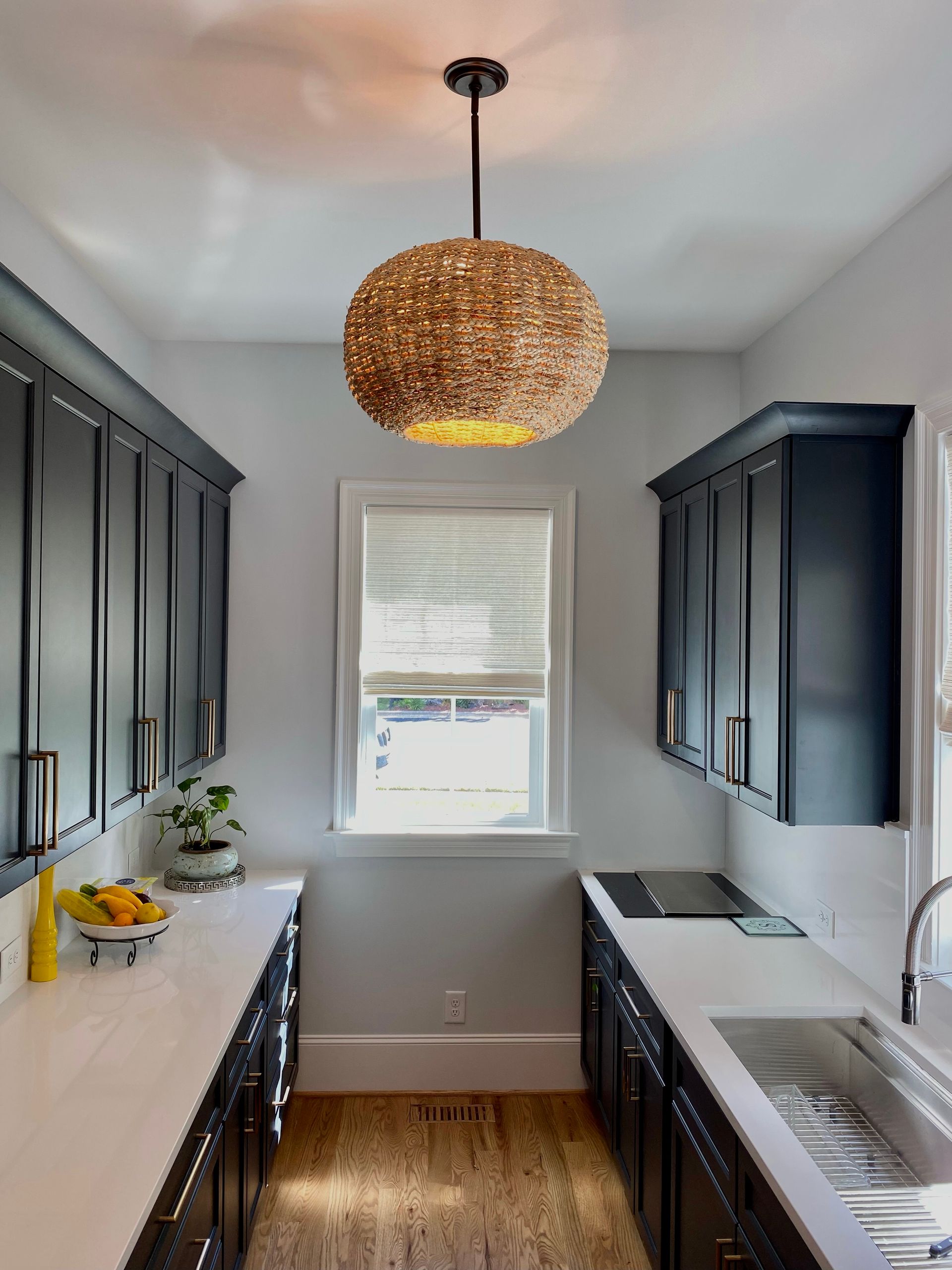Narrow kitchen with dark cabinets, white countertops, and a woven pendant light.