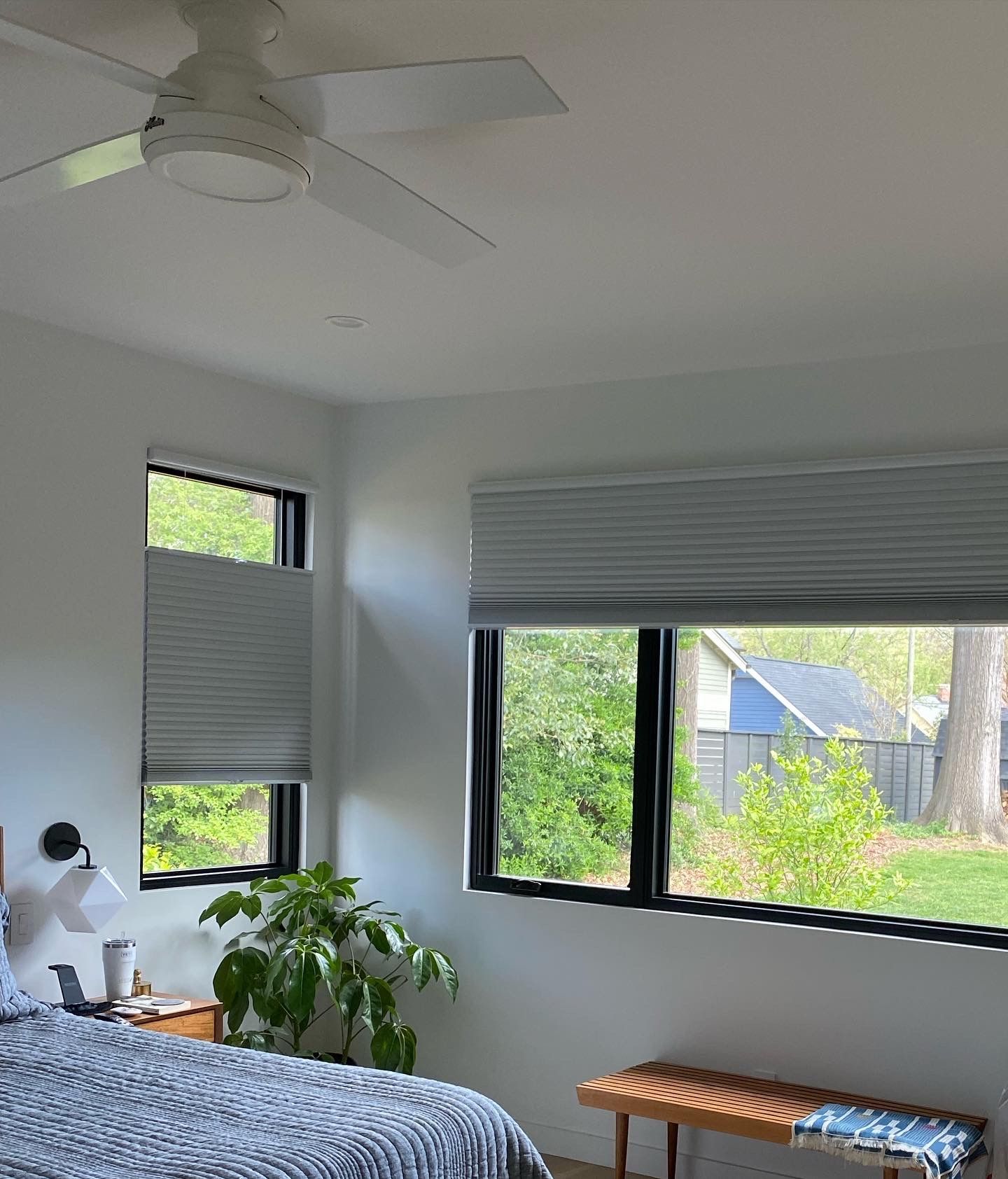 Bedroom with windows, blinds, a ceiling fan, and a bench; plants and trees visible outside.