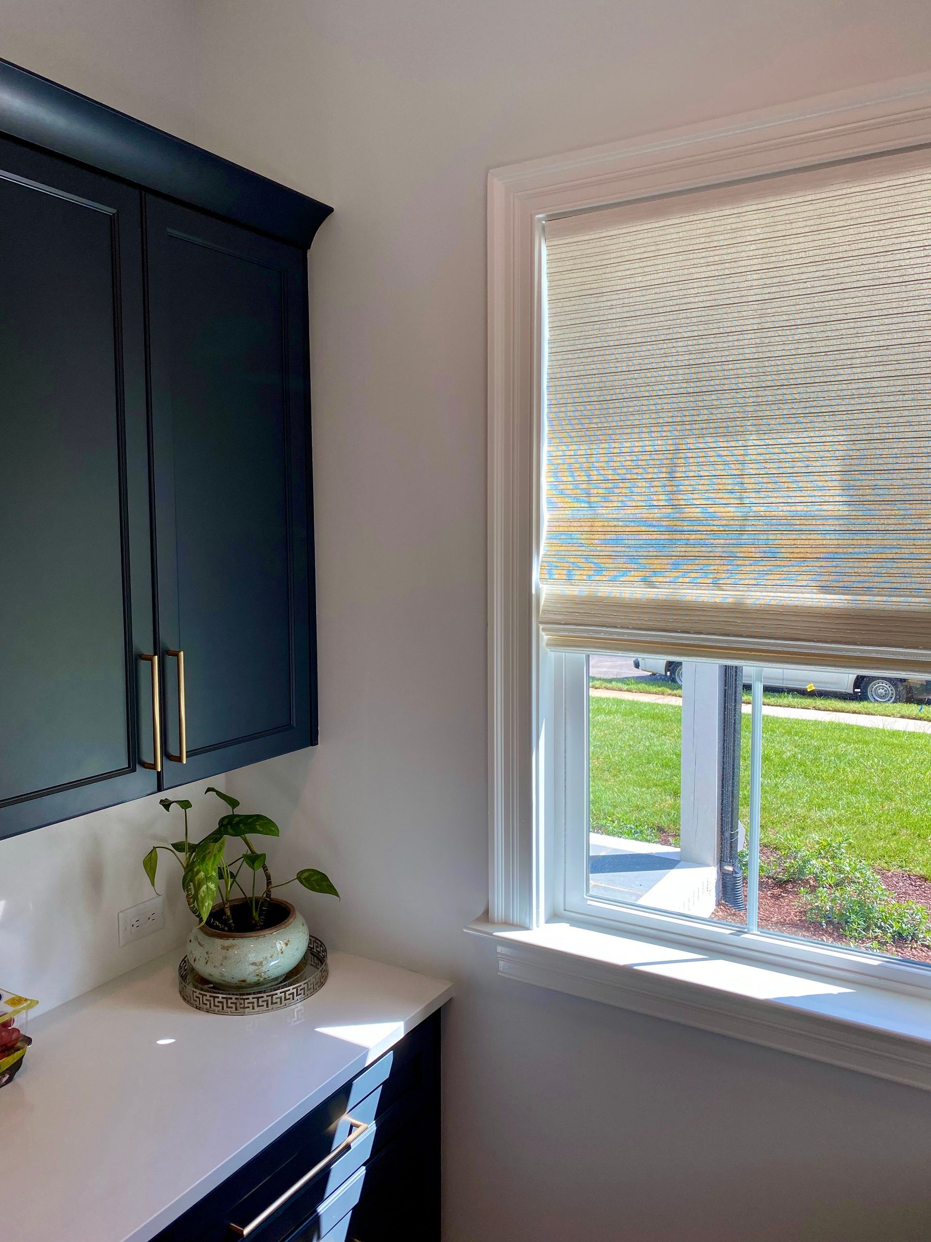 Dark blue cabinets with a white countertop beside a window with a patterned shade, and greenery.