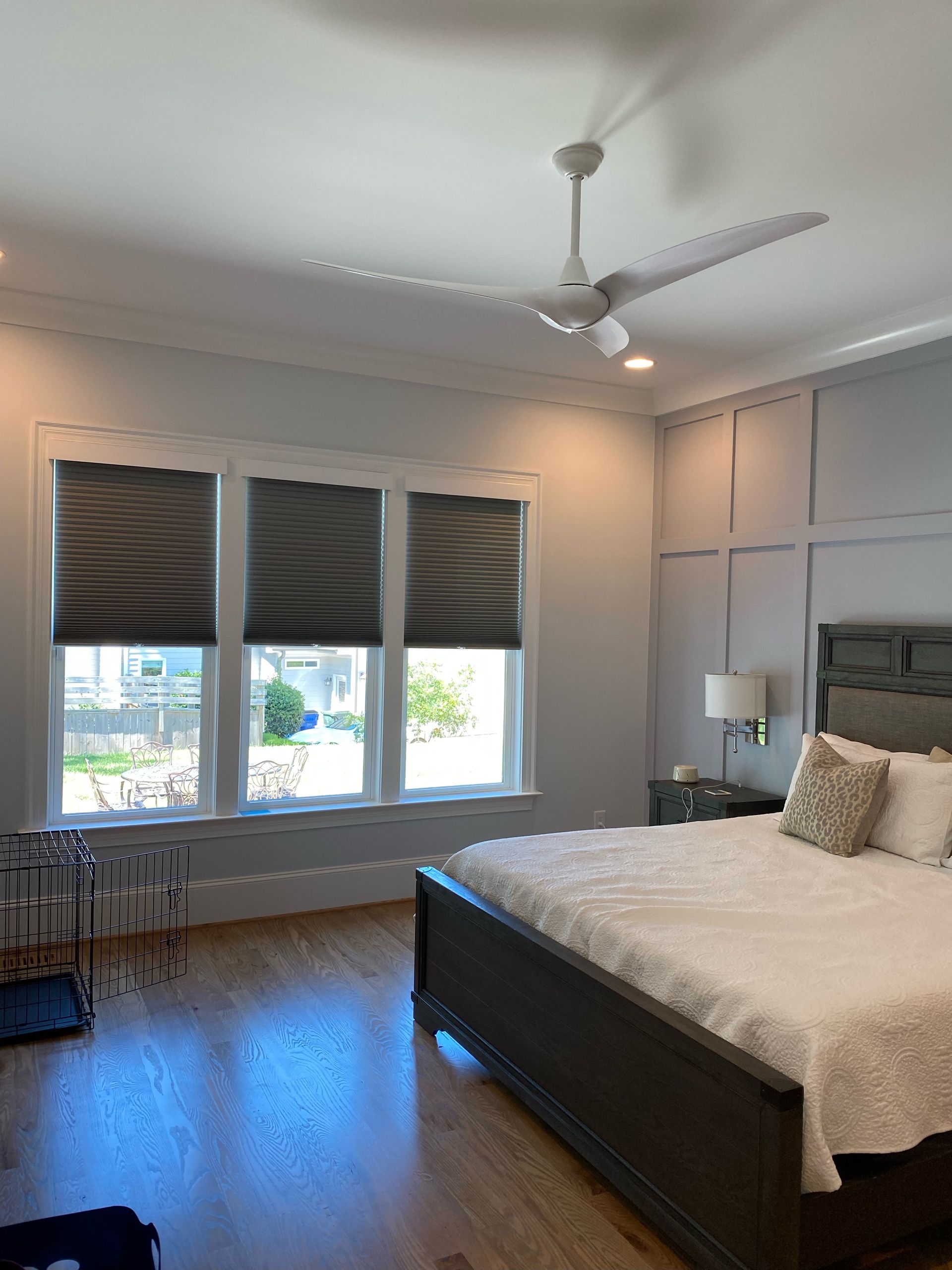 Bedroom with three windows, dark shades, bed, nightstand, and ceiling fan. Wooden floor, gray walls.