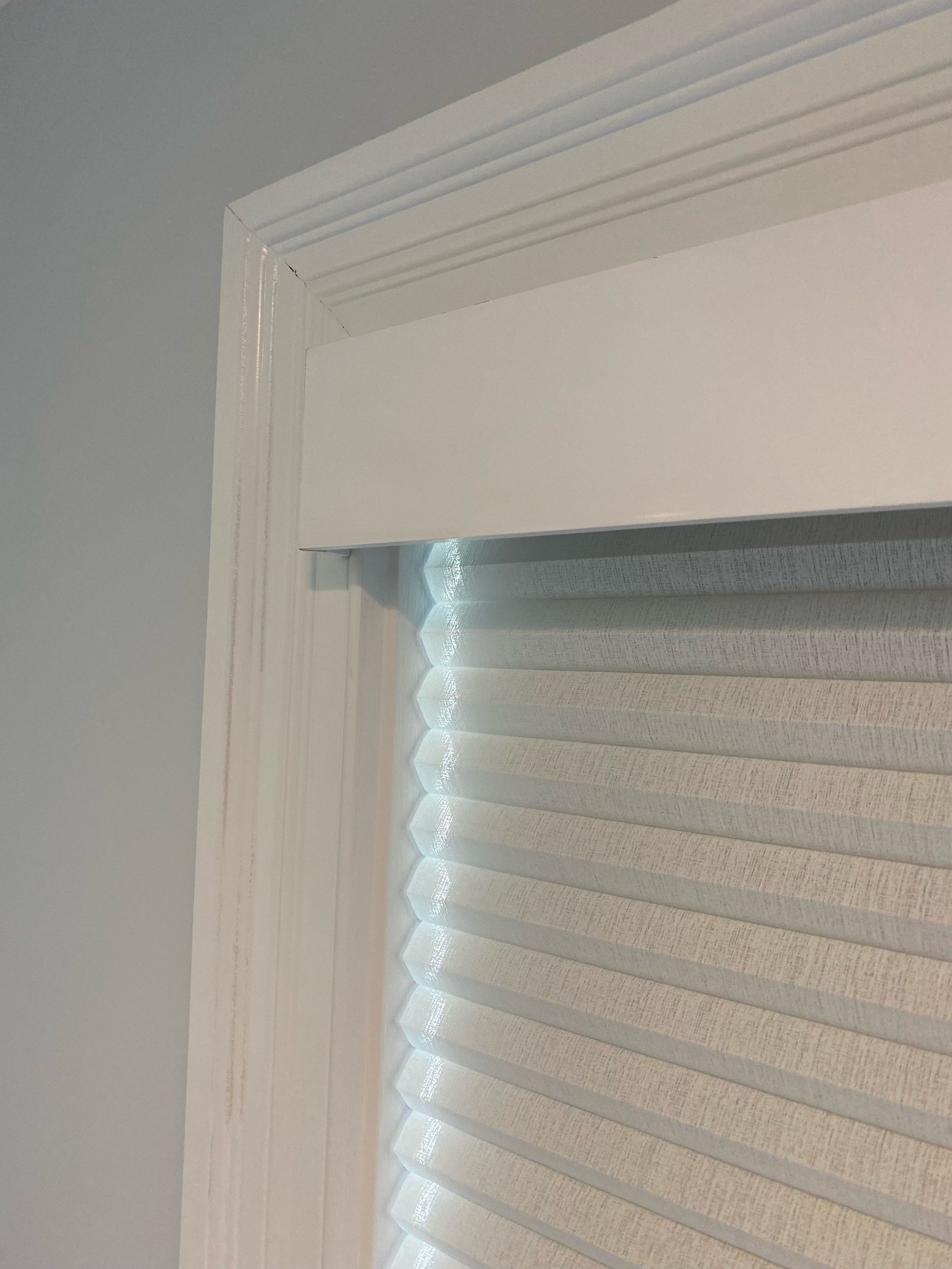 White window frame with a light-colored pleated blind, against a white wall.