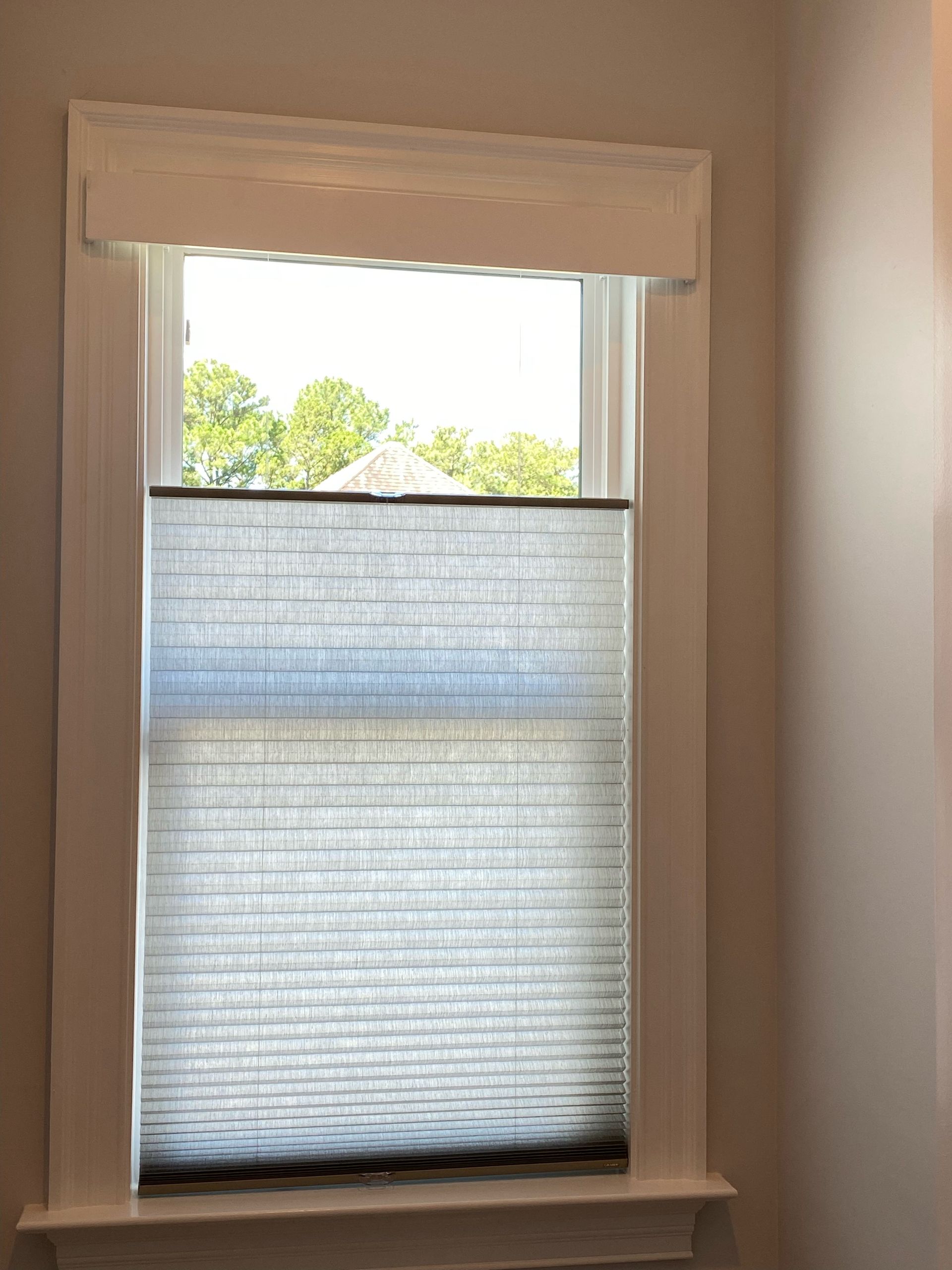 Window with light-filtering cellular shade, framed by white trim and a decorative valance, looking out at trees.