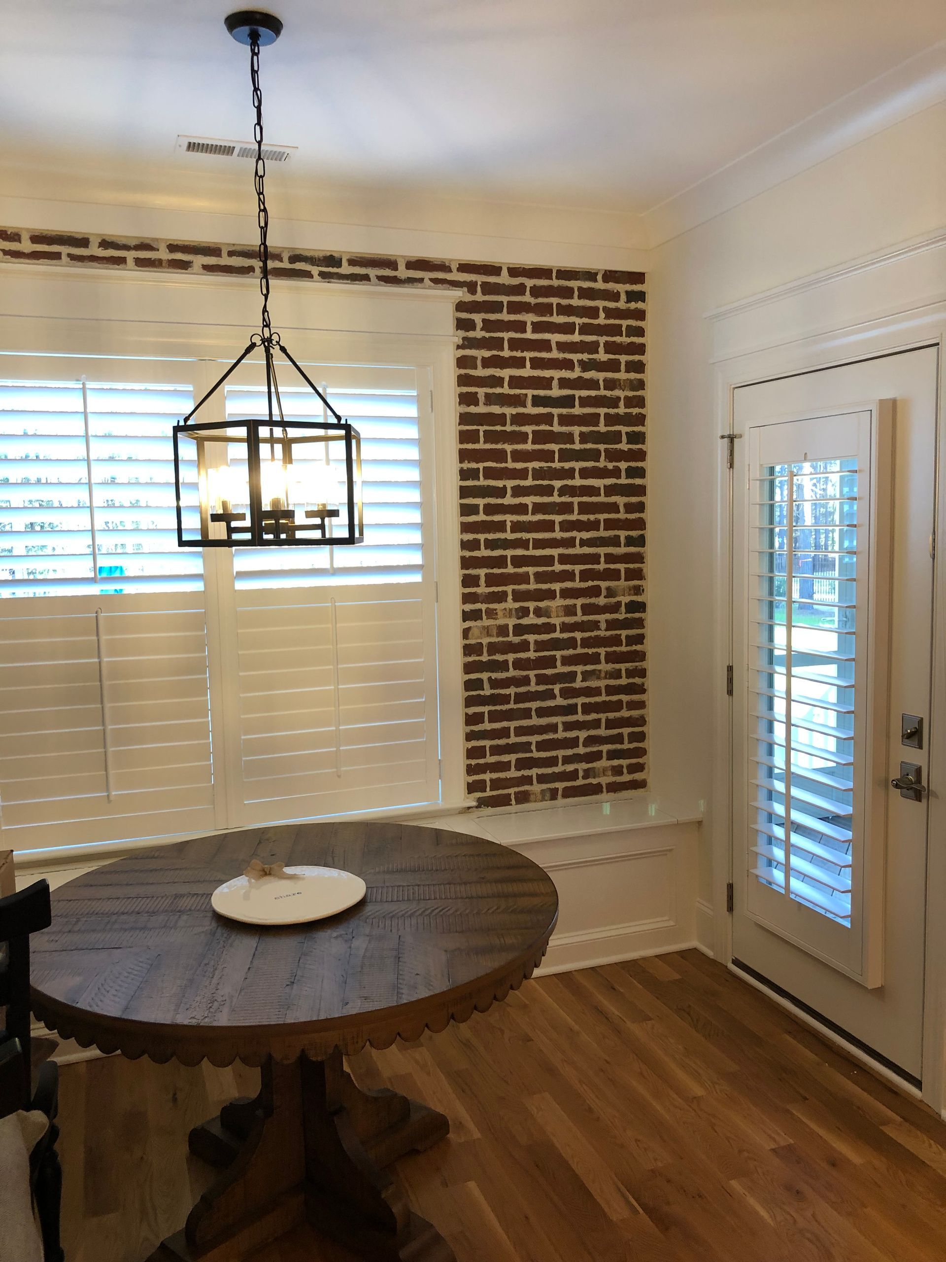 Dining area with round wooden table, brick accent wall, window with shutters, and chandelier.
