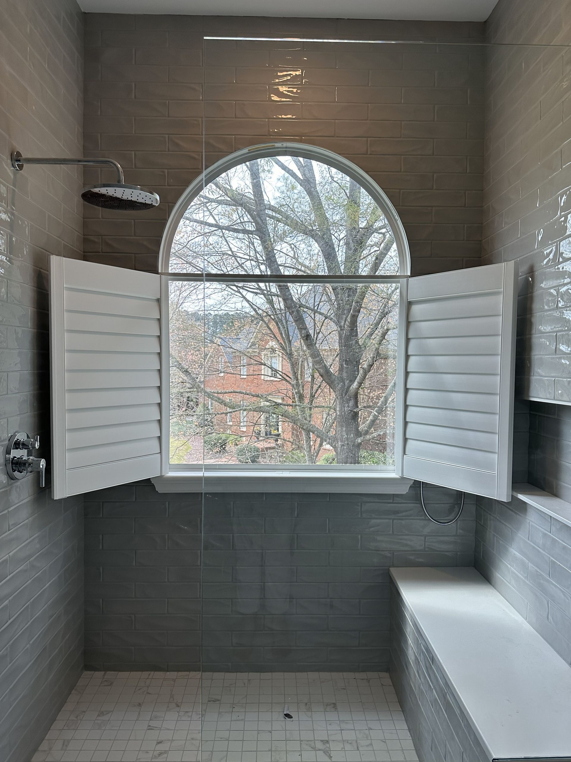 Bathroom with gray tile, open shutters, and a view of trees. Includes a showerhead, bench, and glass door.