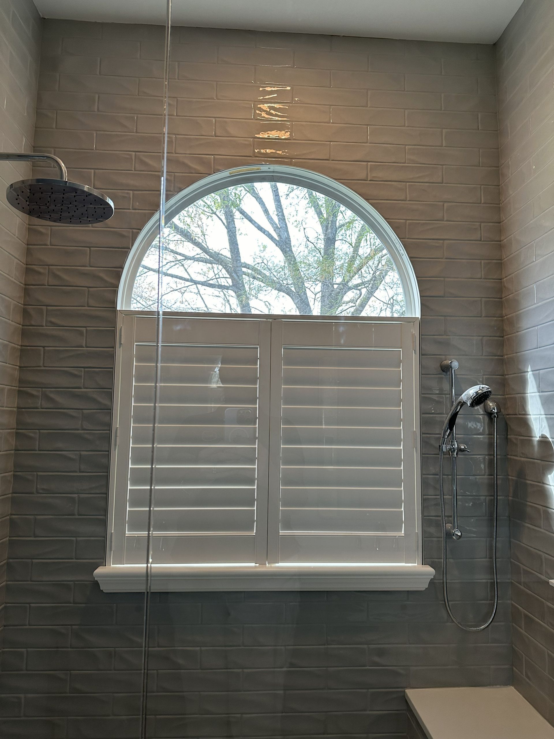 Shower with arched window, white shutters, rain shower head, and gray textured tile.