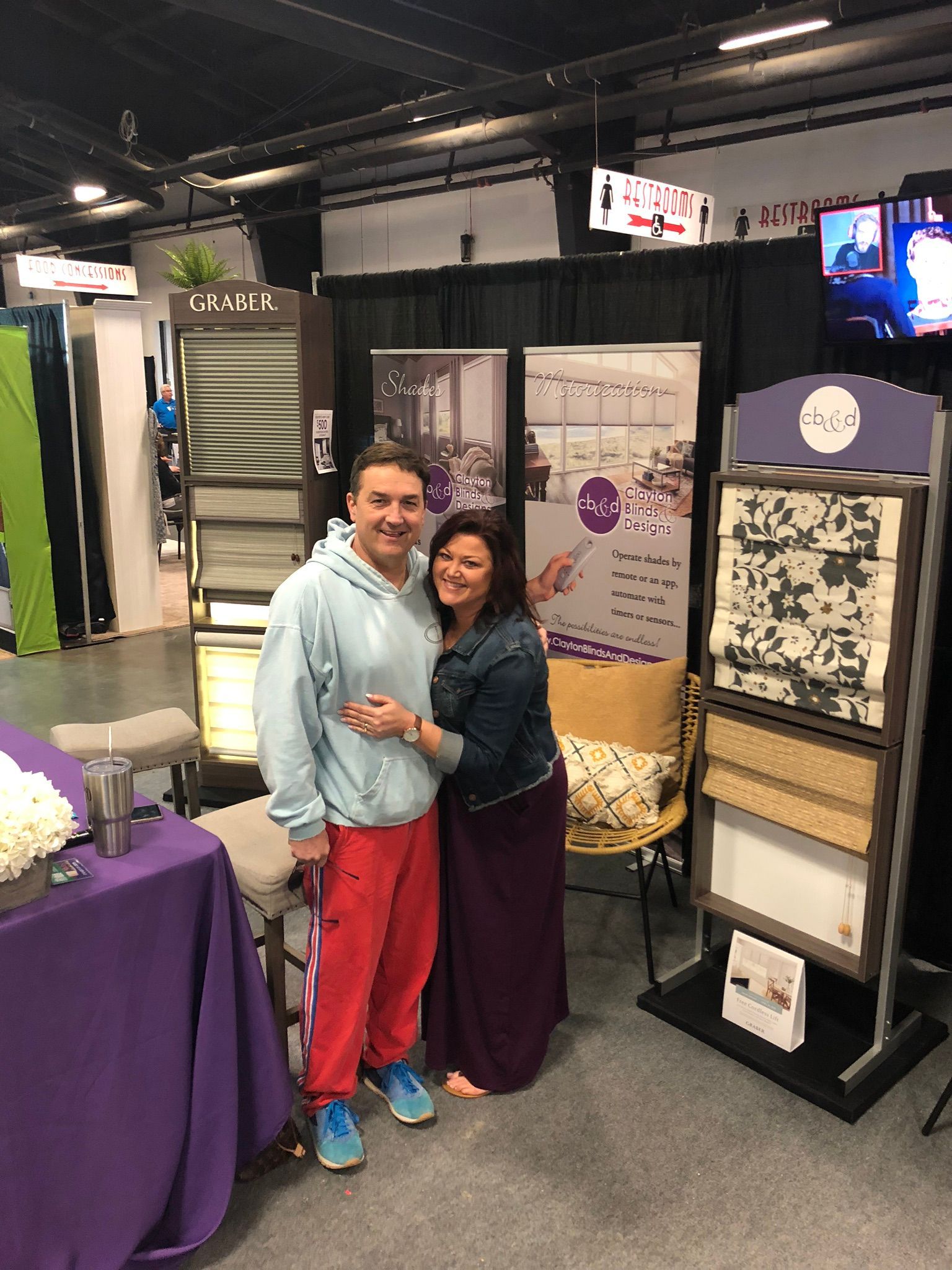 Two people pose at a booth, smiling. Man in red pants, woman in long skirt, both embracing. Booth with display boards and materials.
