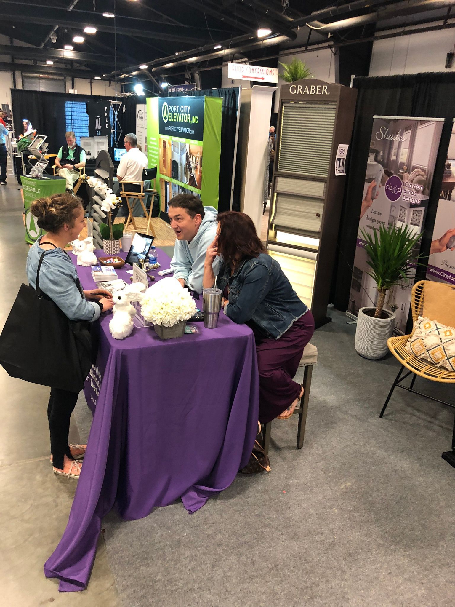 Three people at a purple-covered table in an expo, talking and smiling. A white stuffed animal and flowers are on the table.