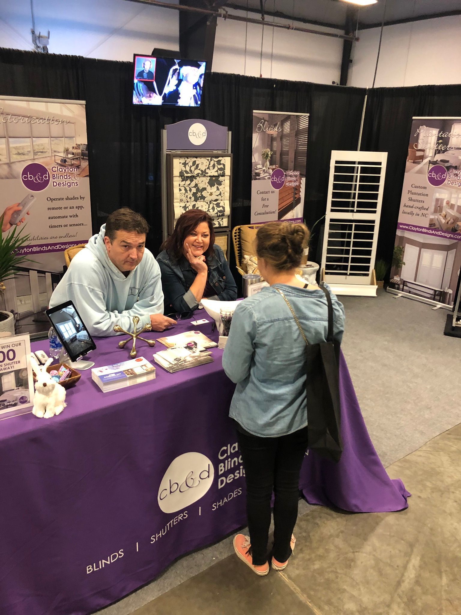 A person at a purple booth with two people behind it. Booth has signs, products, and a small TV.