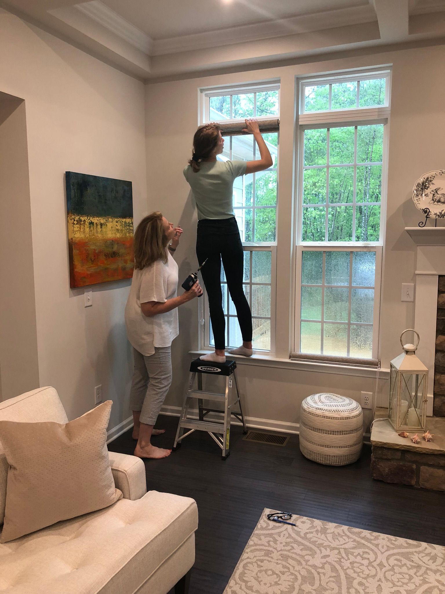 Two people installing window film in a living room. One stands on a stool, working on the window; the other holds something.