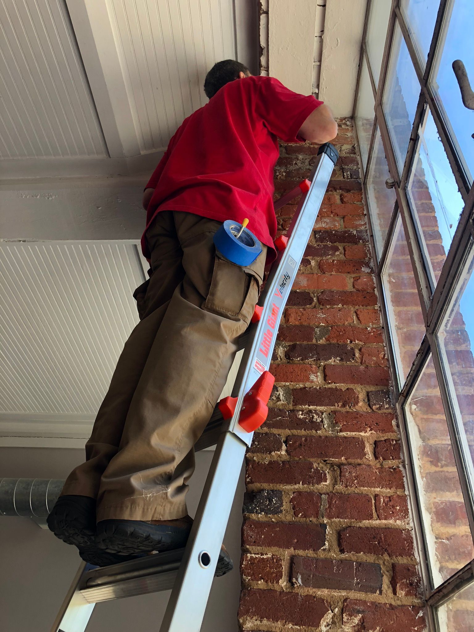 Person on a ladder, working on a building's trim near a brick wall and window.