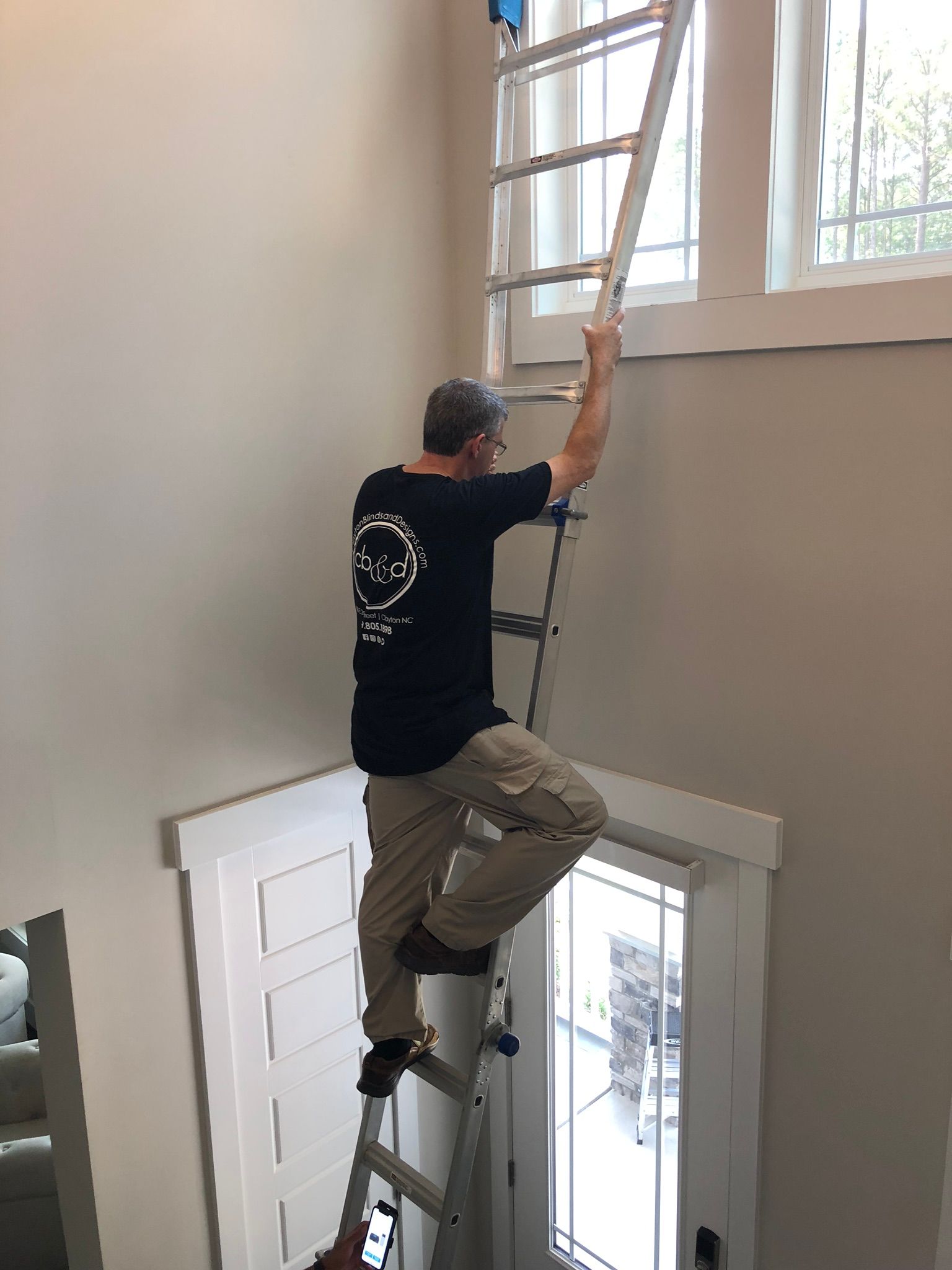 Man on ladder reaching towards a window. Inside a building, beige walls, white trim.
