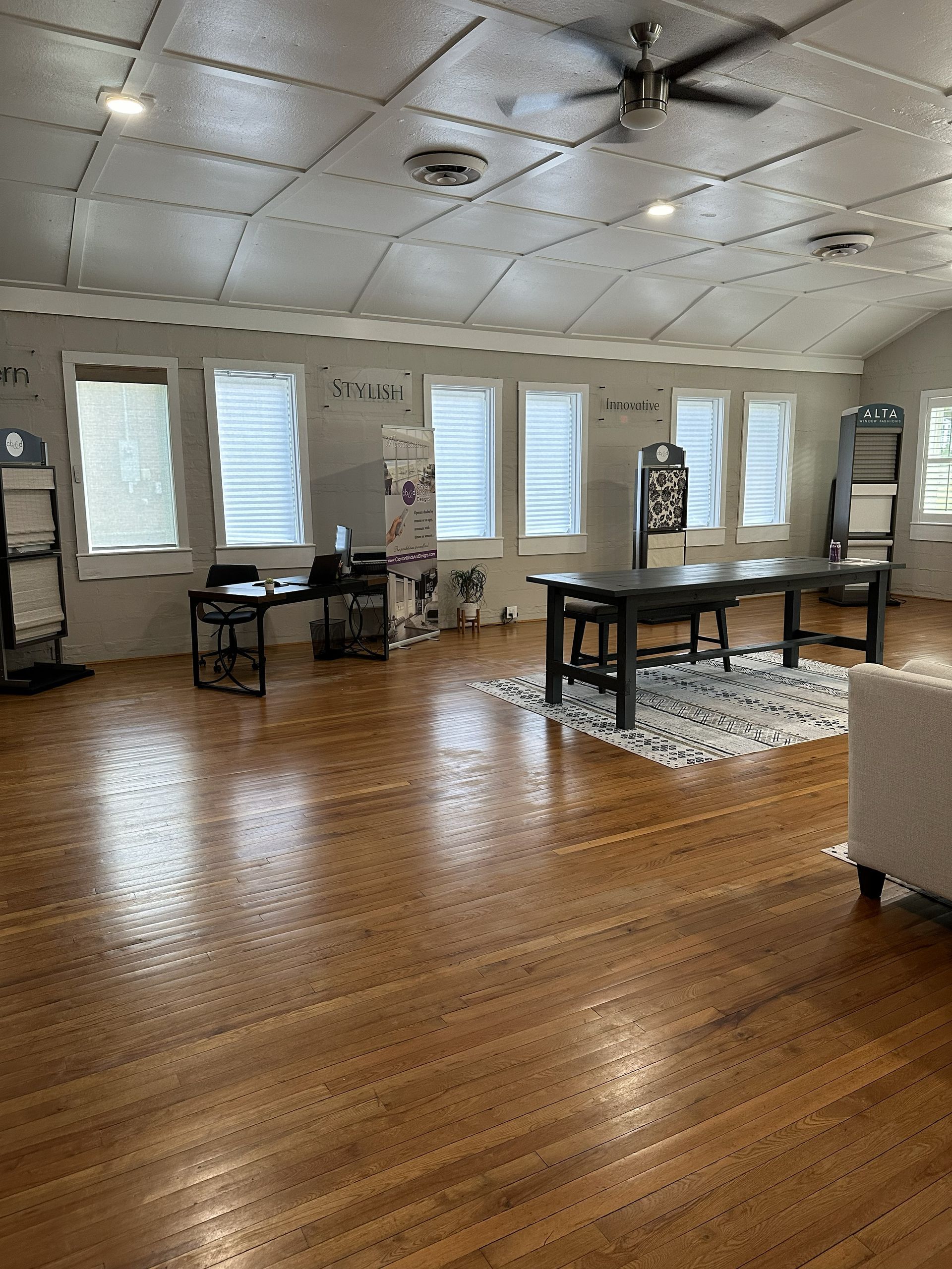 Spacious room with wood floors, several windows, dark tables, and a ceiling fan.