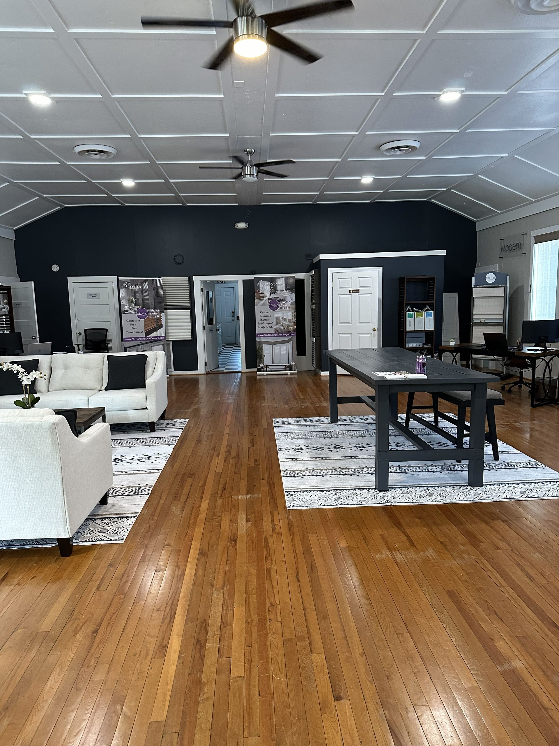 Interior showroom with hardwood floors, white sofas, tables, dark accent wall, and ceiling fans.