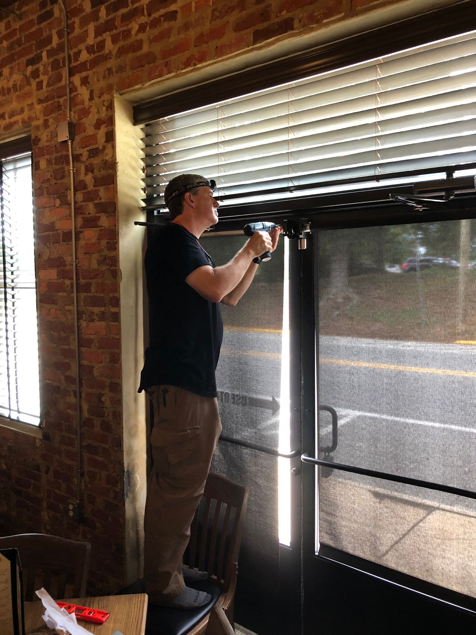 Man on chair repairs a rolling shutter door with a drill. Inside a brick building near a window.