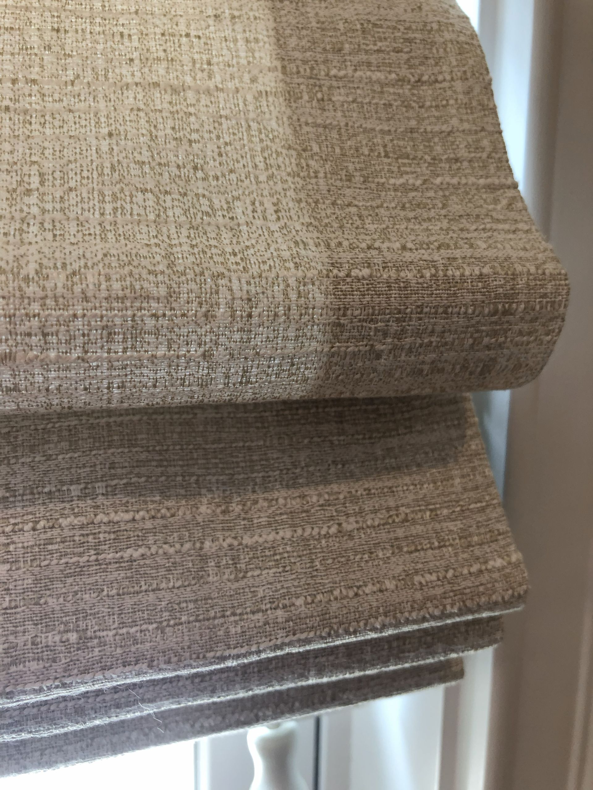 Close-up of beige Roman shade; two folds visible. Textured fabric in front of white window.