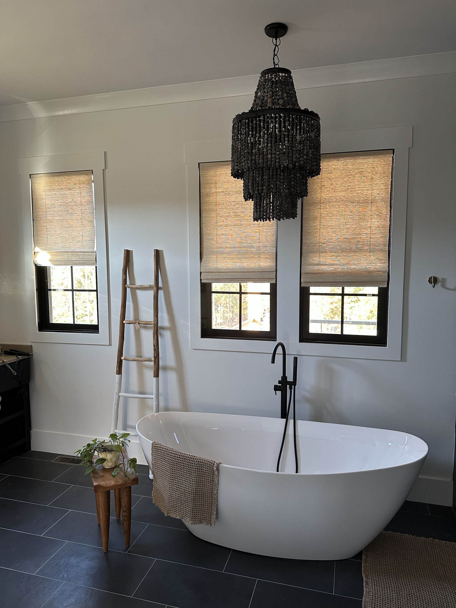 Bathroom with white tub, black chandelier, three windows with shades, wooden ladder.