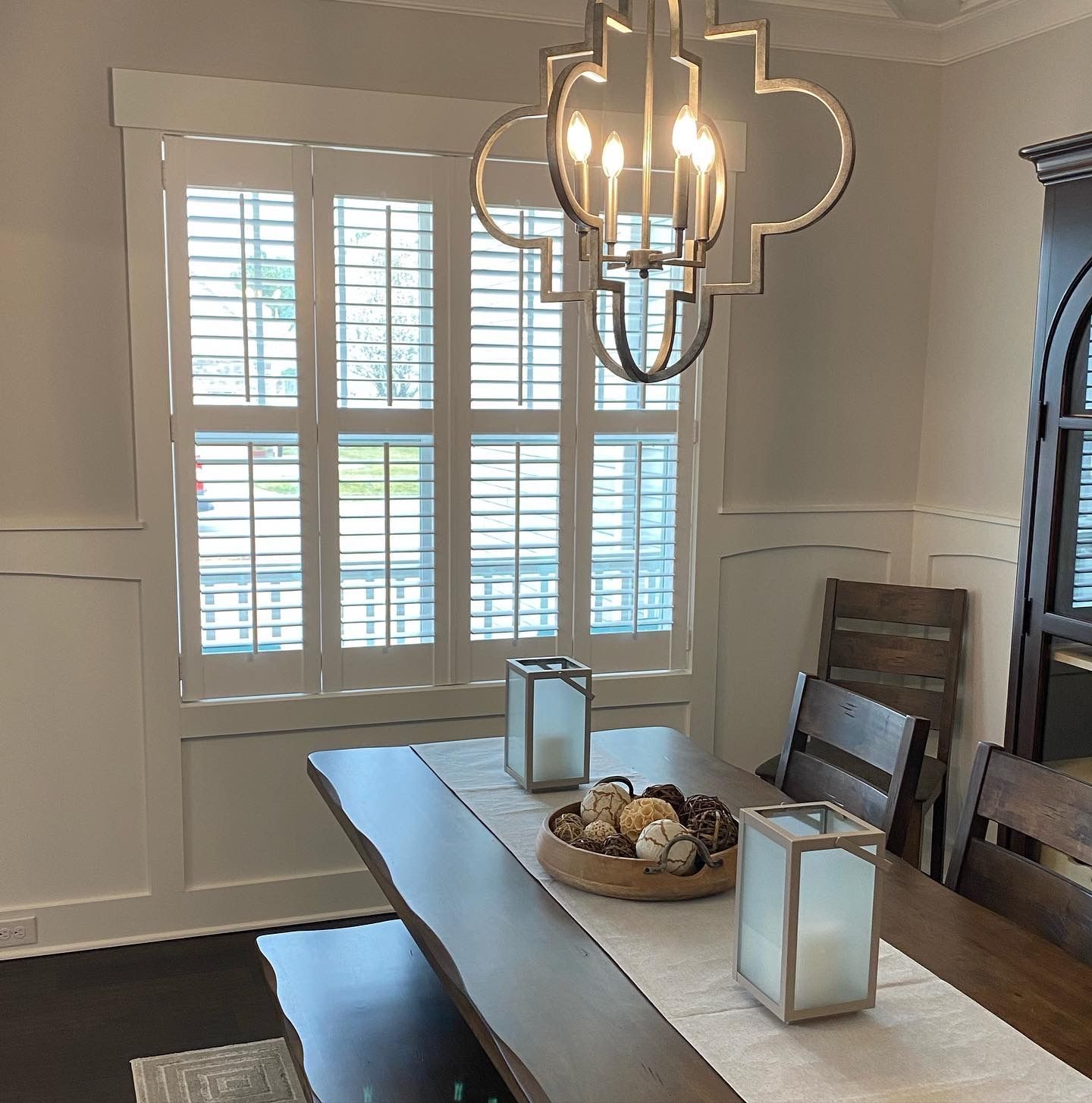 Dining room with wooden table, white shutters, and a hanging chandelier.