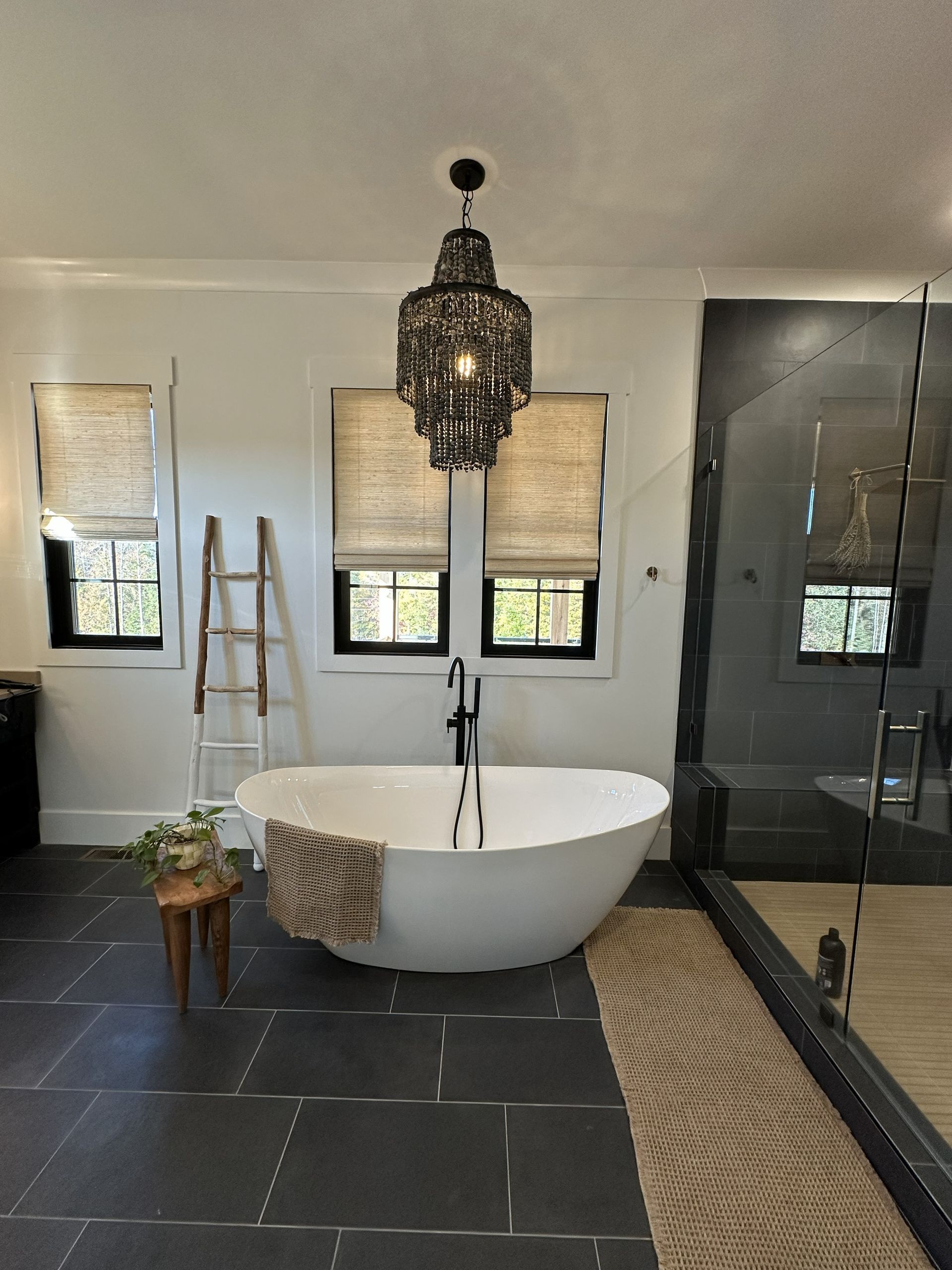 Elegant bathroom with a black chandelier above a white soaking tub. Black tile floor, windows with blinds.