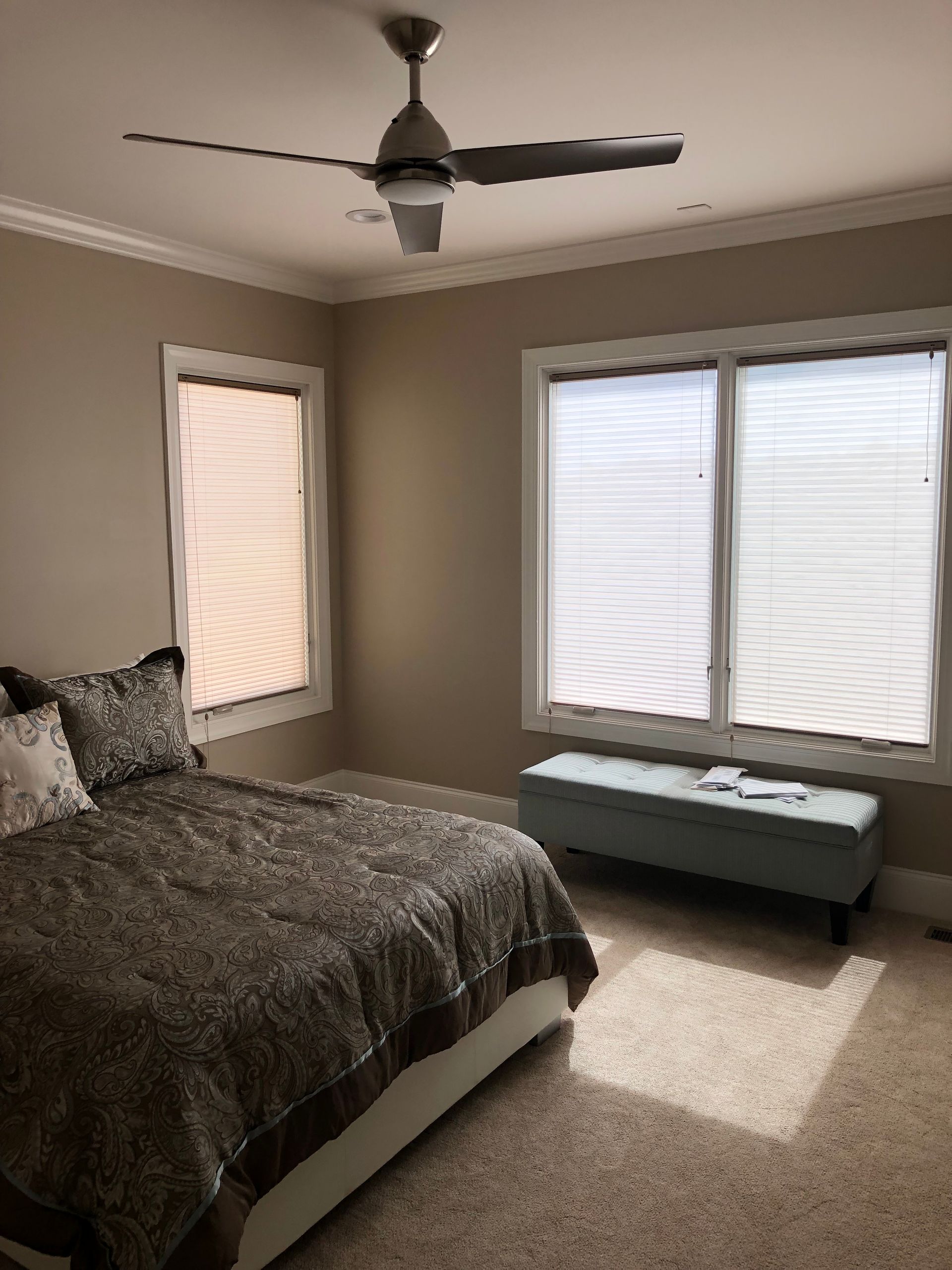 Bedroom with bed, windows, ceiling fan, and blue storage bench. Beige walls, neutral-toned carpet.