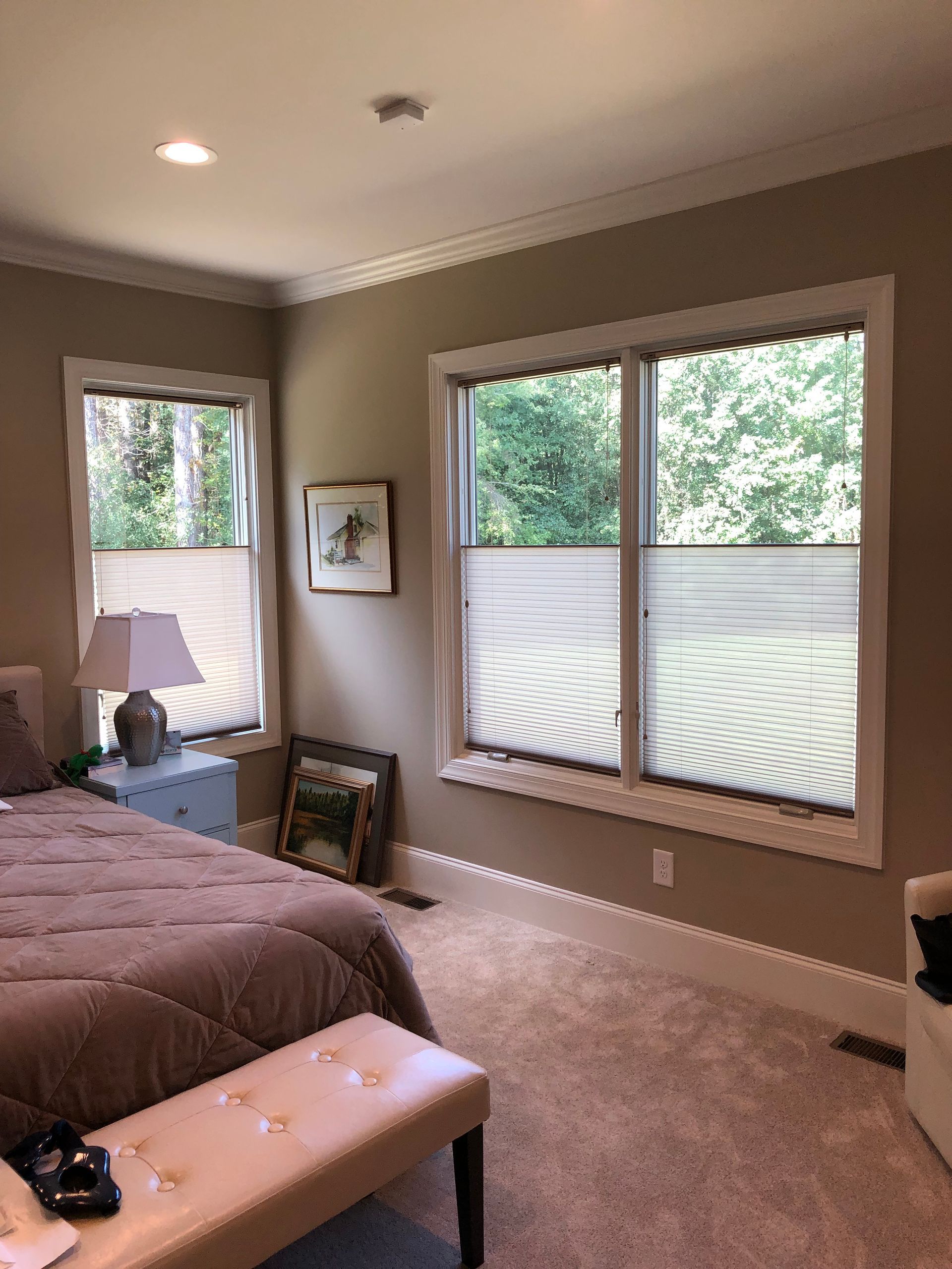 Bedroom with windows and beige walls. Bed with brown comforter, window coverings, artwork, and bench.
