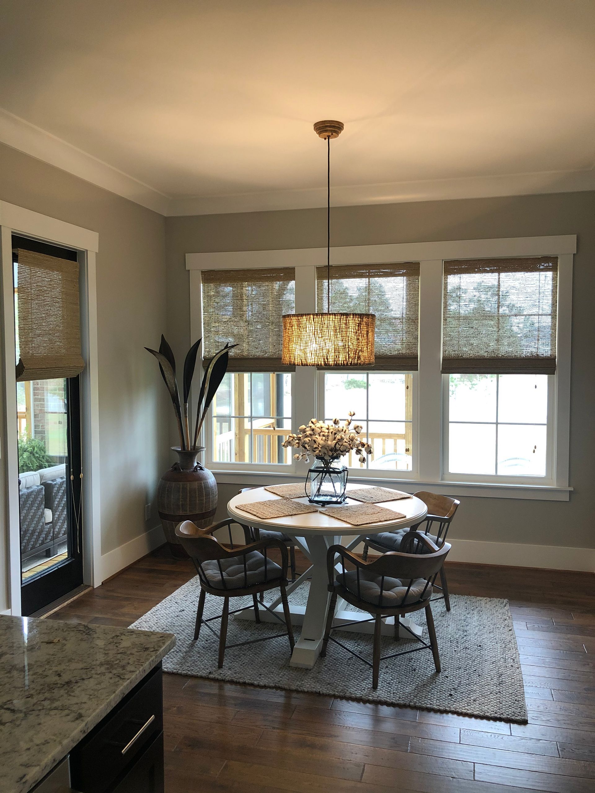 Dining room with round table, chairs, and woven shades. A vase holds tall, dark branches.