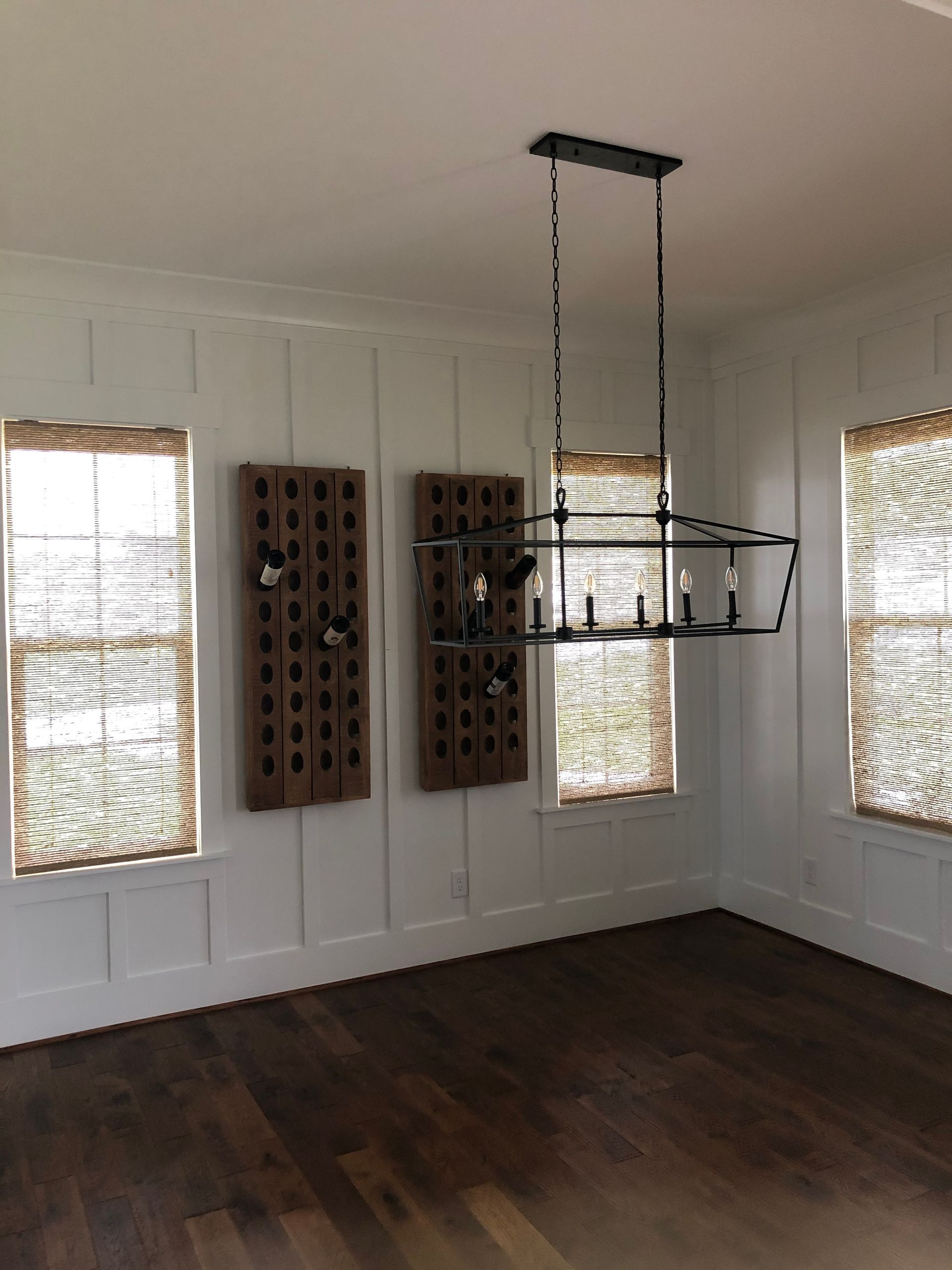 Empty dining room with dark wood floor, white walls with wainscoting, woven window shades, and a black chandelier.