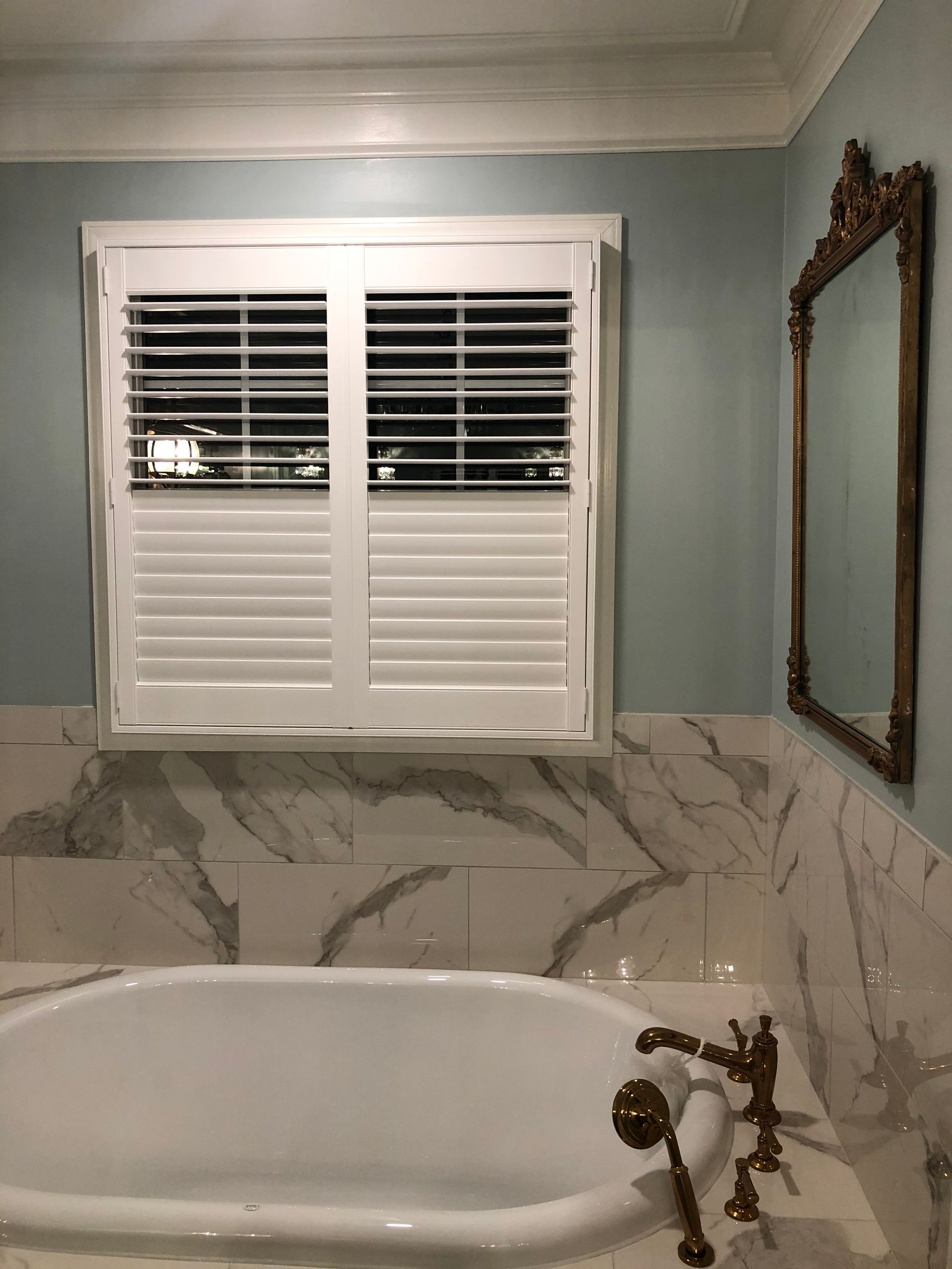Bathroom with a white bathtub, shuttered window, and ornate mirror. Soft blue walls and marble accents.