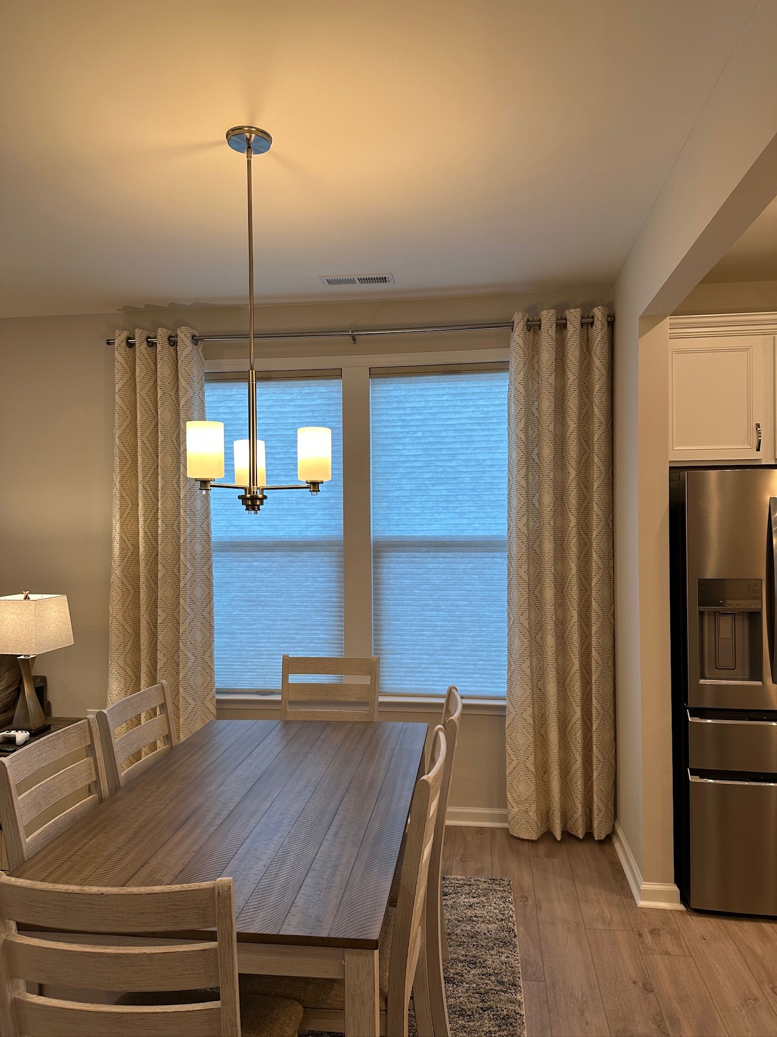 Dining room with long wooden table, chandelier, curtains, and windows with blinds. A rug is beneath the table.
