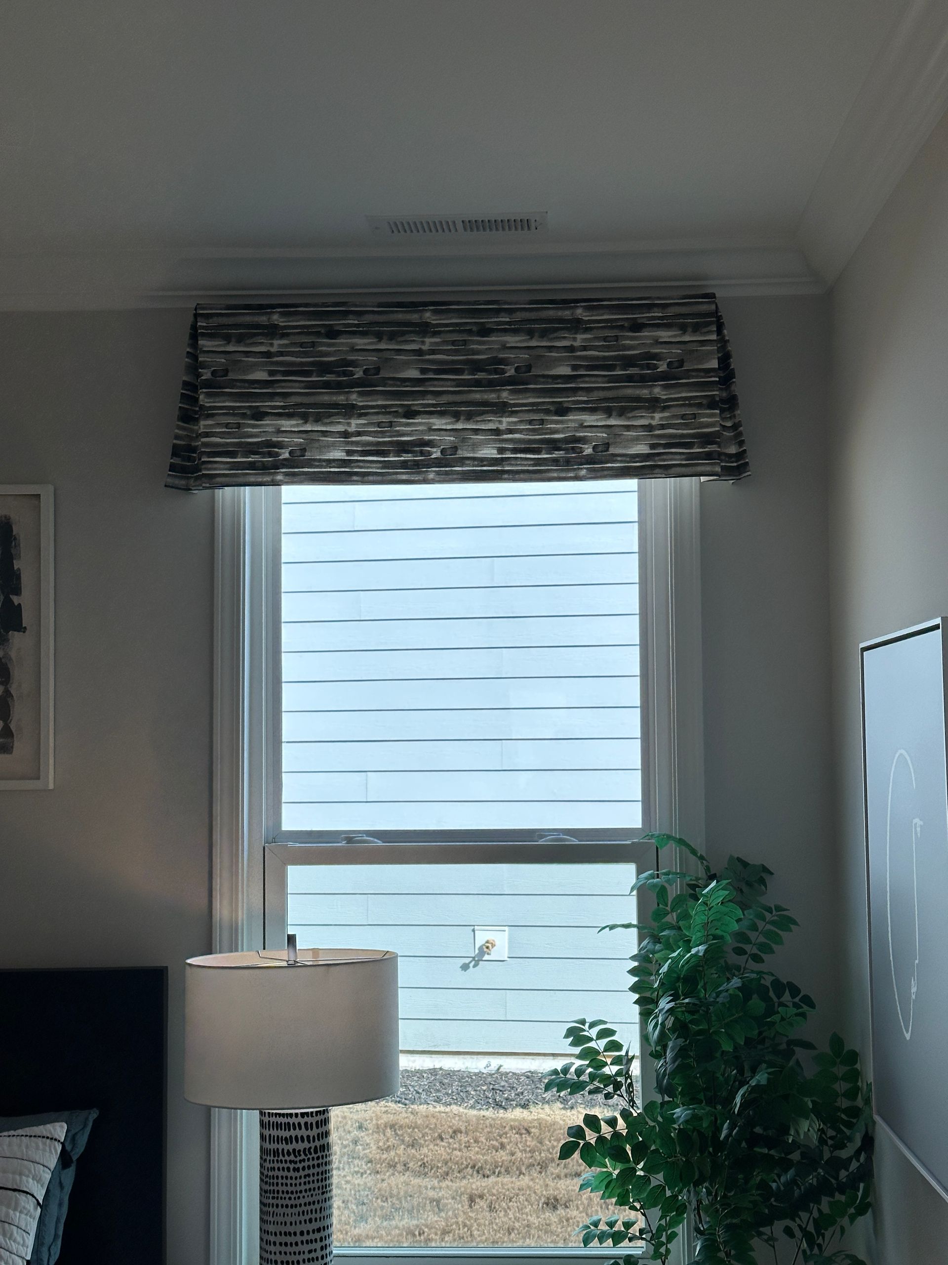 Window with a dark patterned valance, white blinds, and a view of an outside landscape.