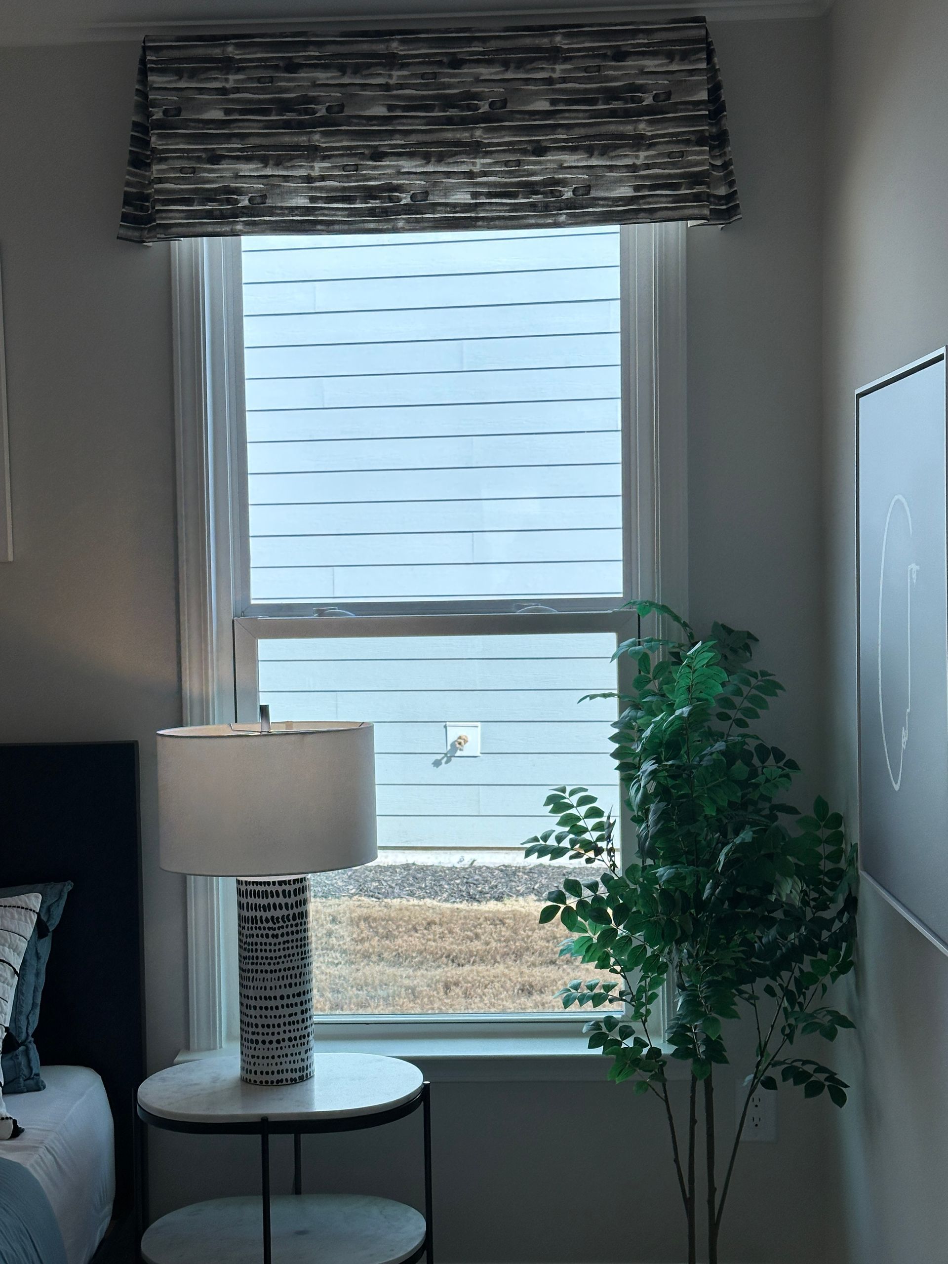 Window with a patterned valance, white blinds, a lamp, and a plant inside a room with neutral walls.