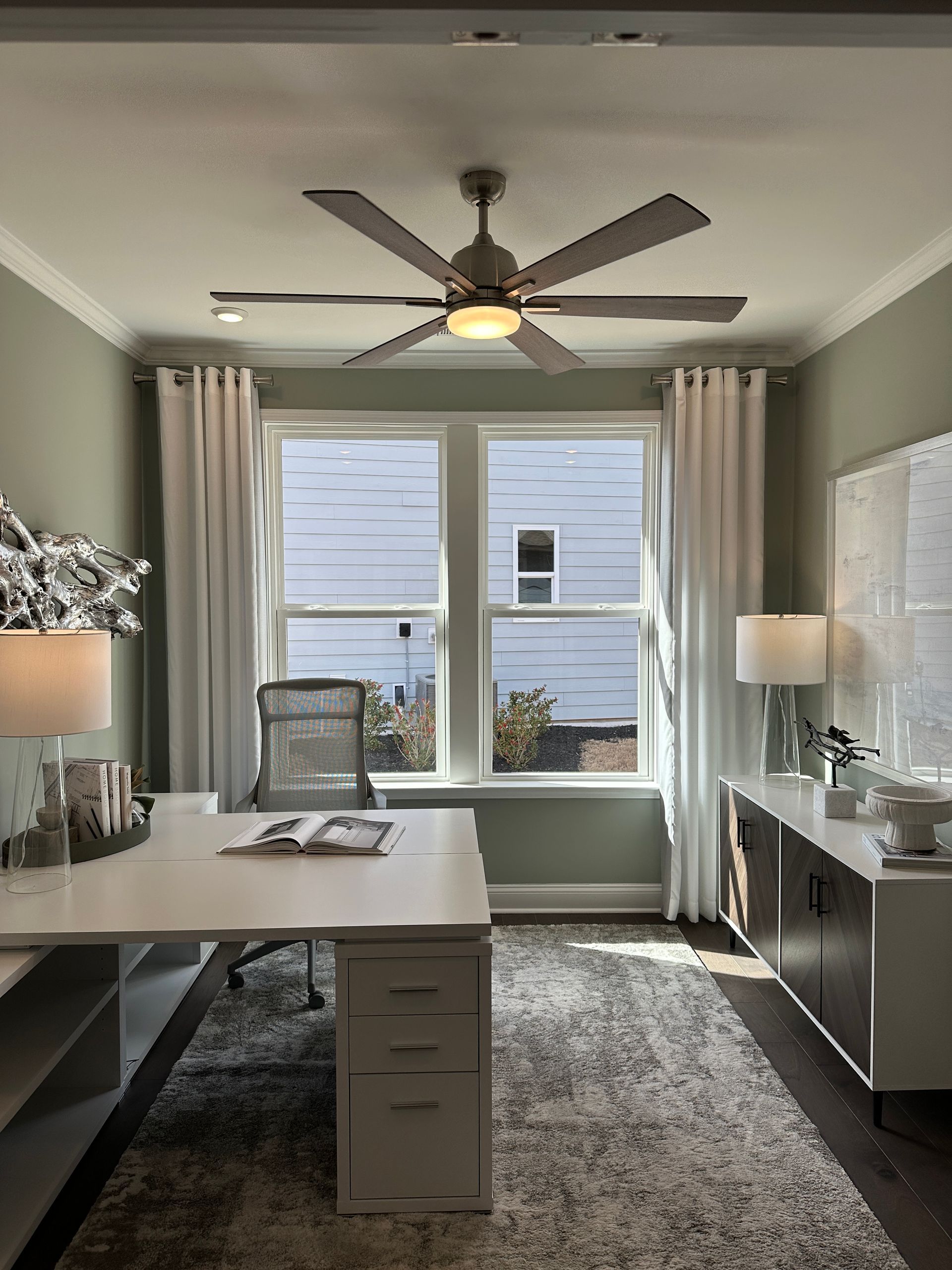 Office space with a white desk, grey rug, windows, and ceiling fan.