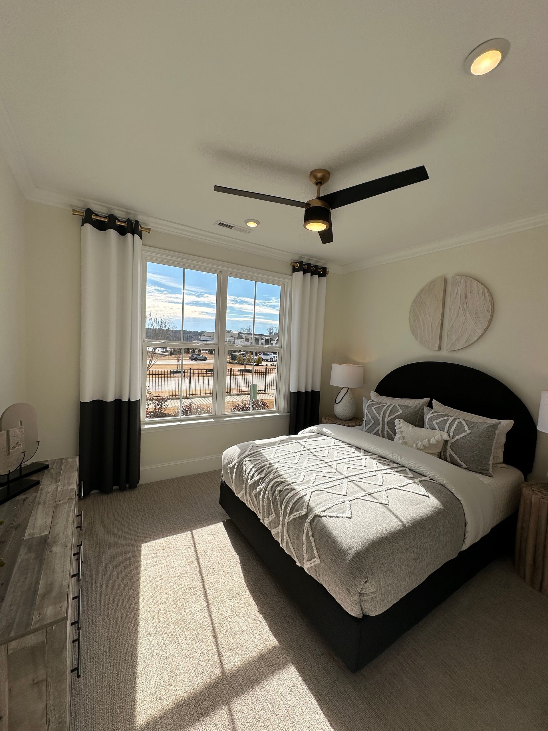 Bedroom with a bed, window, dresser, and ceiling fan. White and black curtains frame the window with a view.