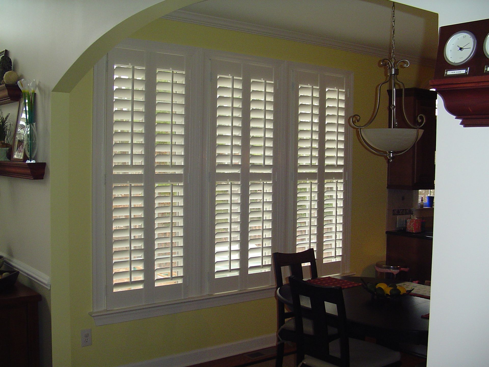 Three white shuttered windows in a room with a dining area. Yellow walls and wood furniture are present.