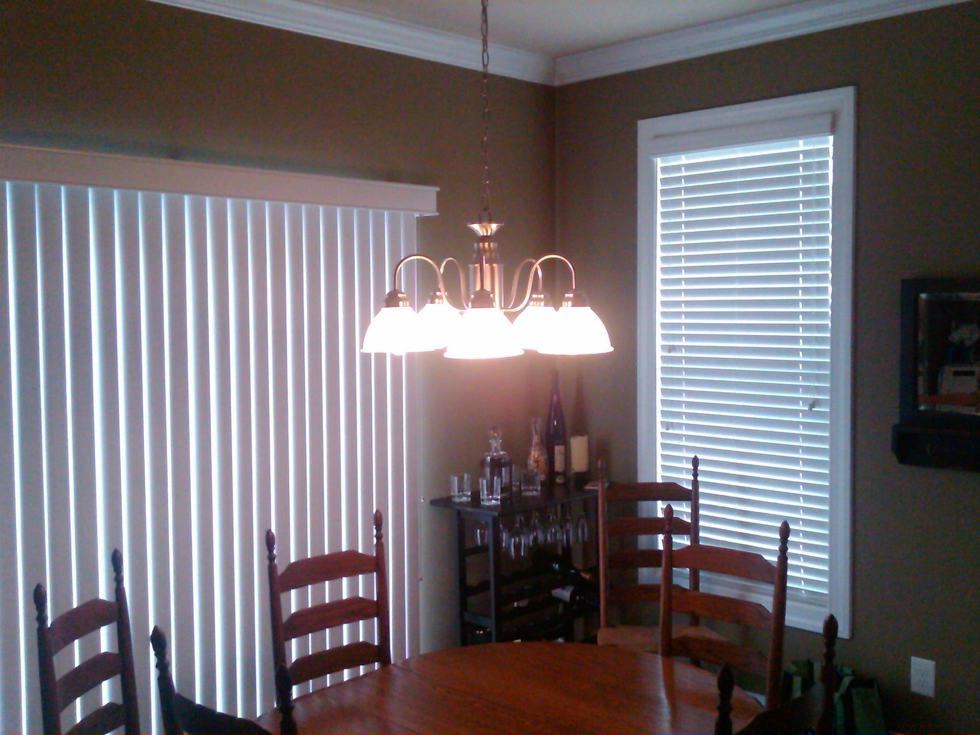 Dining room with table, chairs, vertical blinds, chandelier, wine rack, and brown walls.
