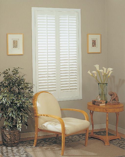 Interior room with a window covered by white shutters, armchair, potted plant, and small table.