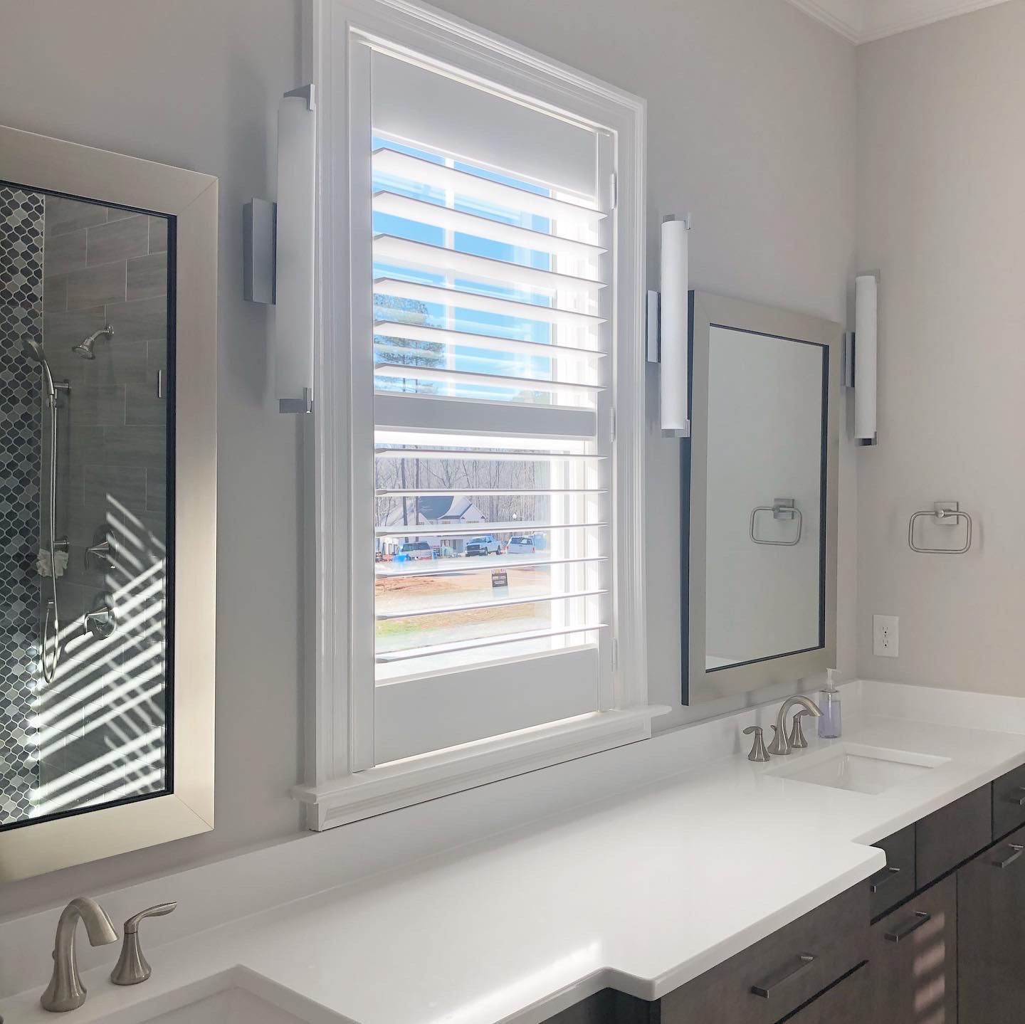 Bathroom with a long white countertop, mirrors, a window with shutters, and two sinks.