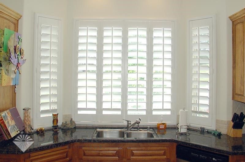 White shuttered kitchen windows above a sink with a dark countertop and wooden cabinets.
