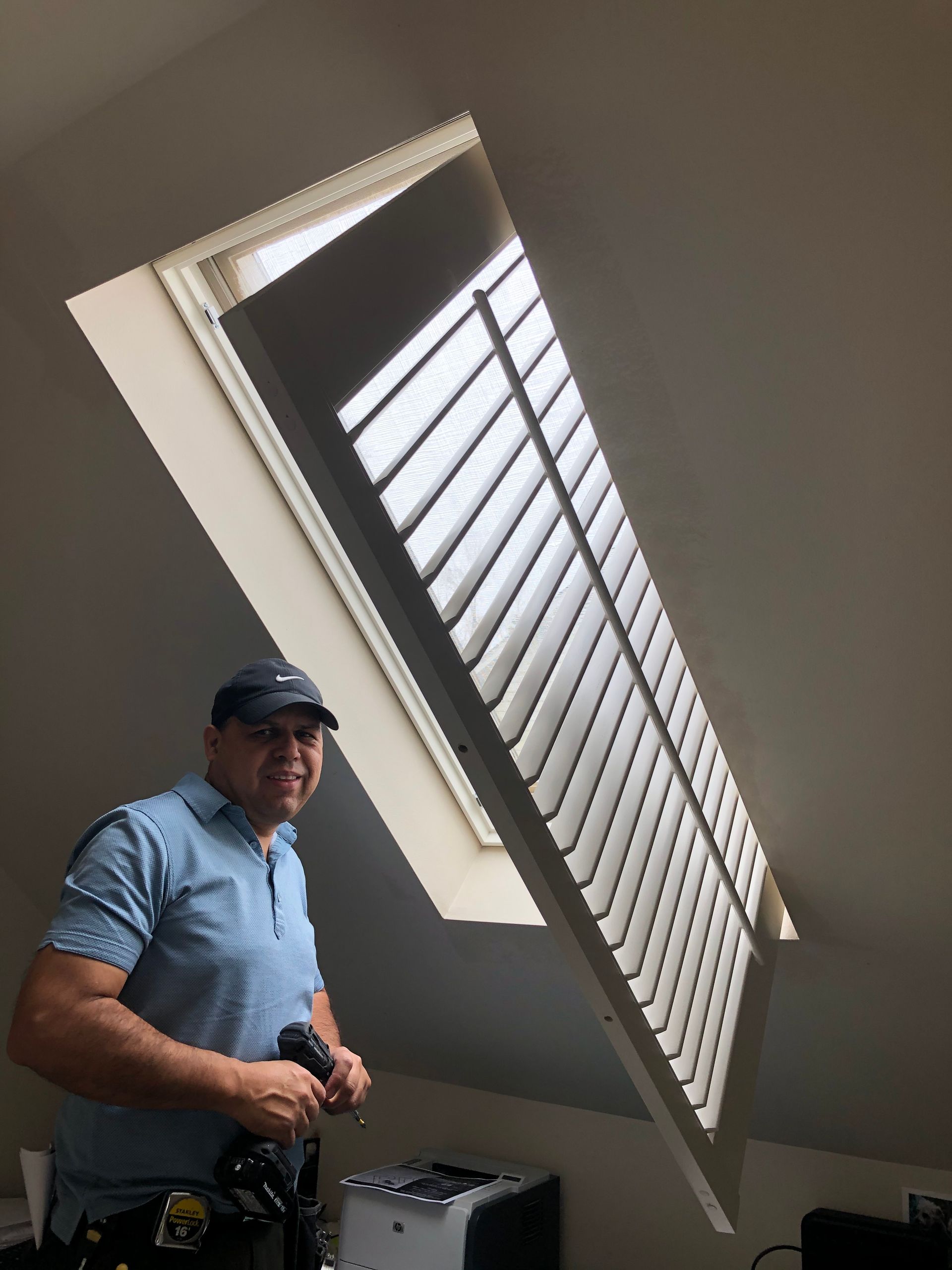 Man installing interior shutters on a slanted skylight. He holds a tool and wears a cap.