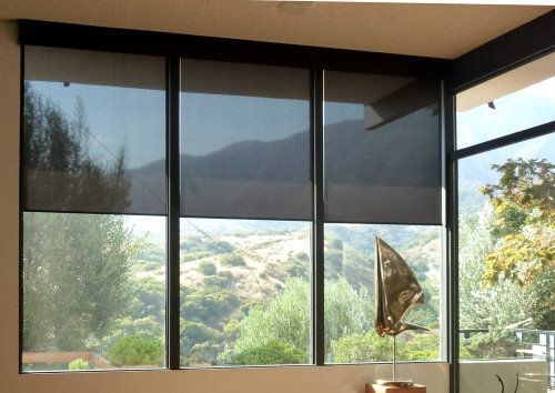 Large windows with dark gray roller shades, overlooking a sunny hillside.