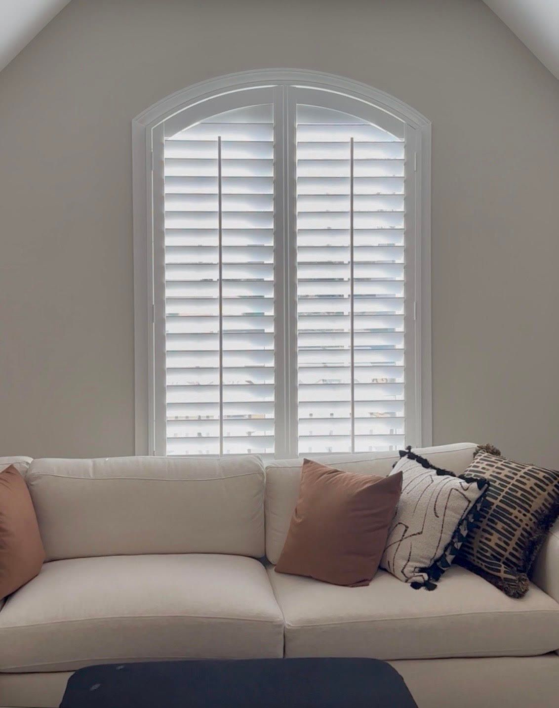 White couch with orange and patterned pillows in front of arched window with white shutters.