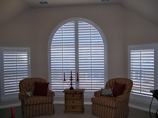 Two upholstered chairs flank a small table with a candelabra, beneath arched window with shutters.