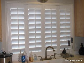 White shutters on a kitchen window, partially open, with a faucet and sink in the foreground.