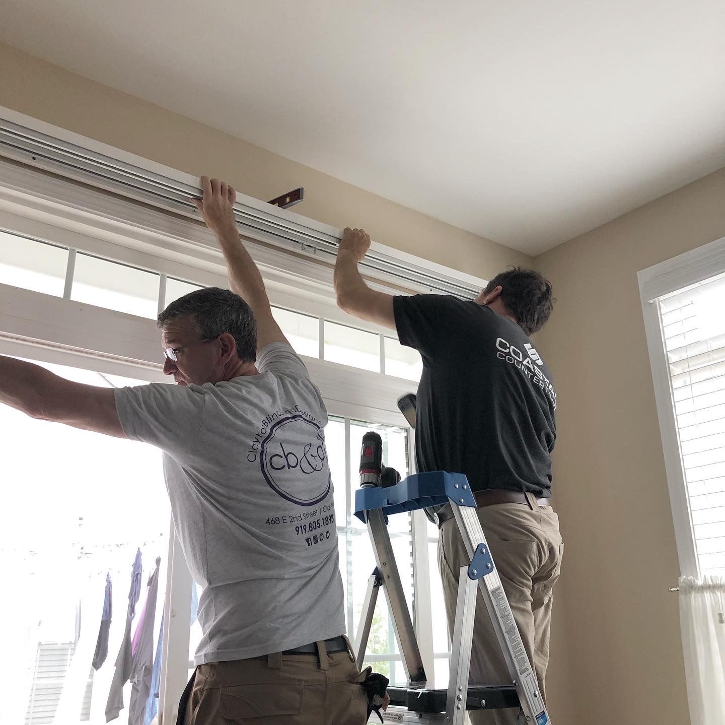 Two men install window blinds. One on a ladder, the other standing near a window. Interior setting.
