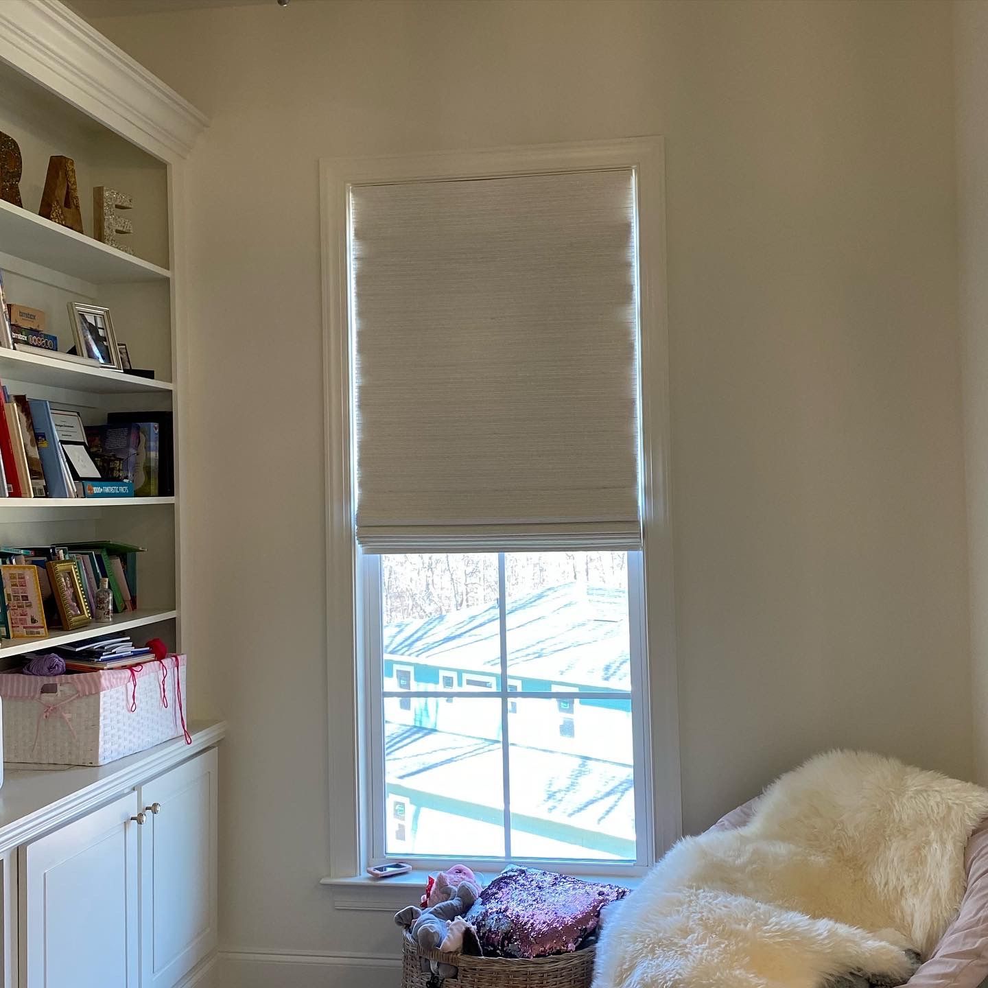 Window with cream-colored shade, natural light, and a white-framed window. Bookshelf on the left, chair on the right.
