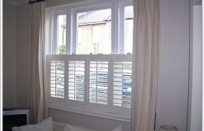 White window with closed shutters, flanked by cream-colored curtains, indoors.