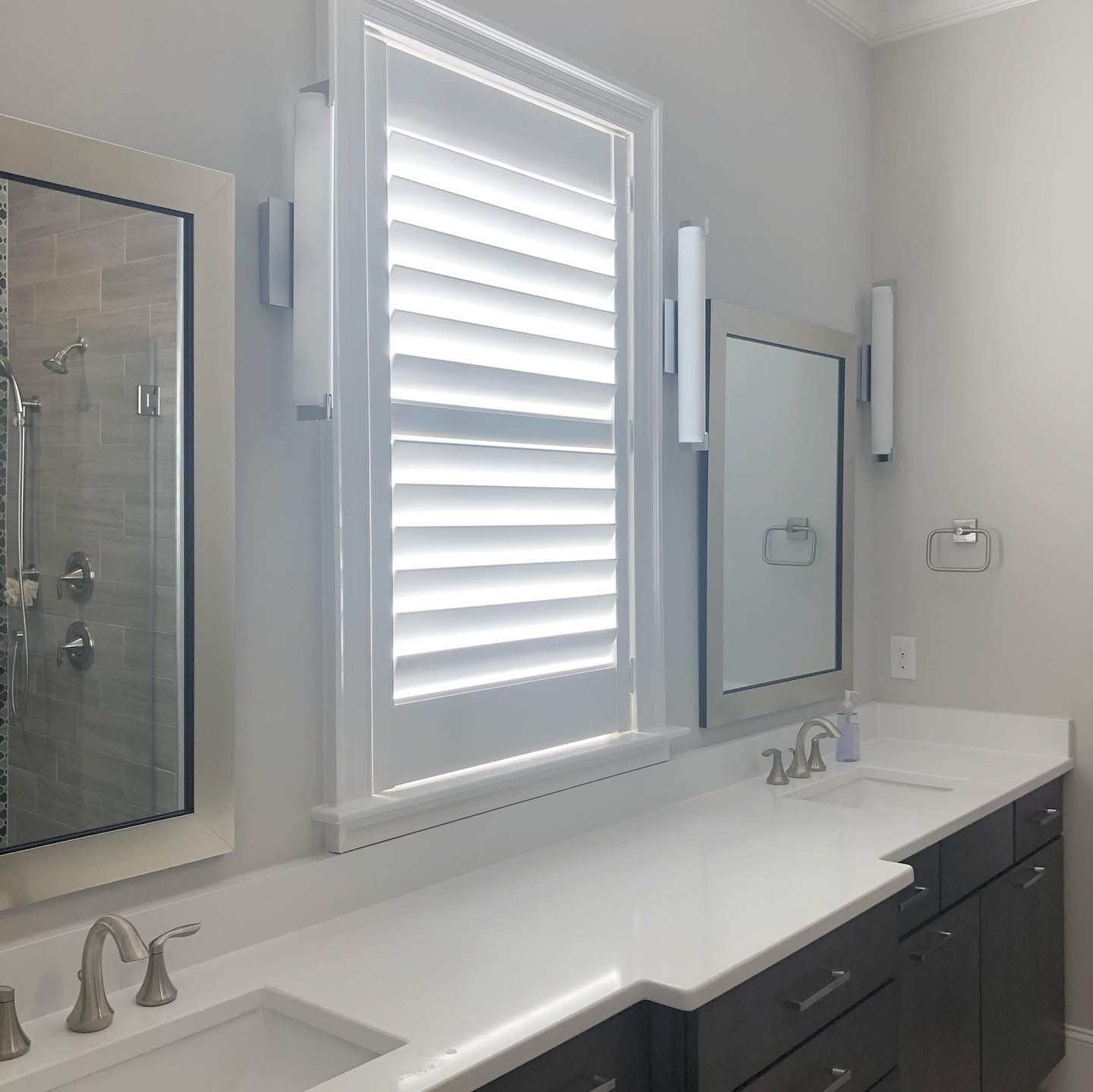 Bathroom with white countertop, dark cabinets, two mirrors, a window with shutters, and brushed nickel fixtures.