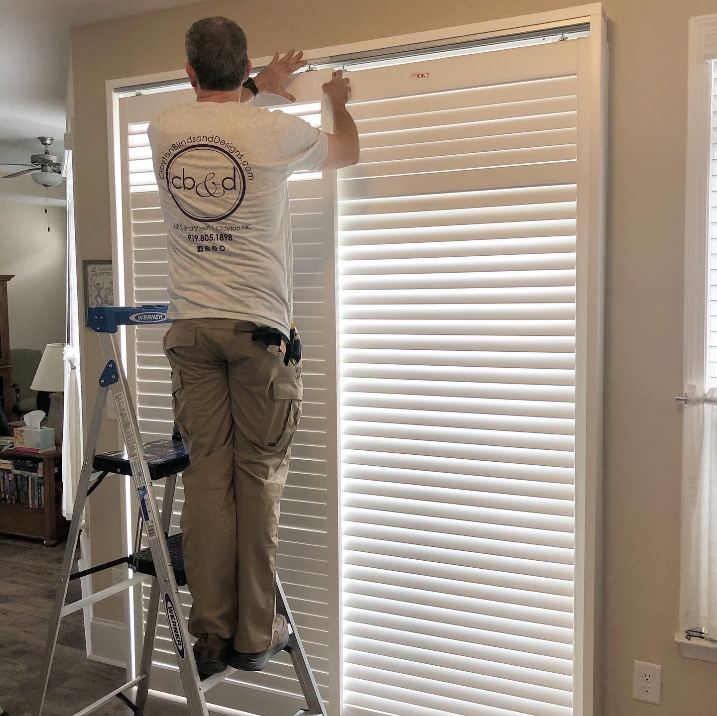 Person on a stepladder installing white shutters on a window. Interior setting.